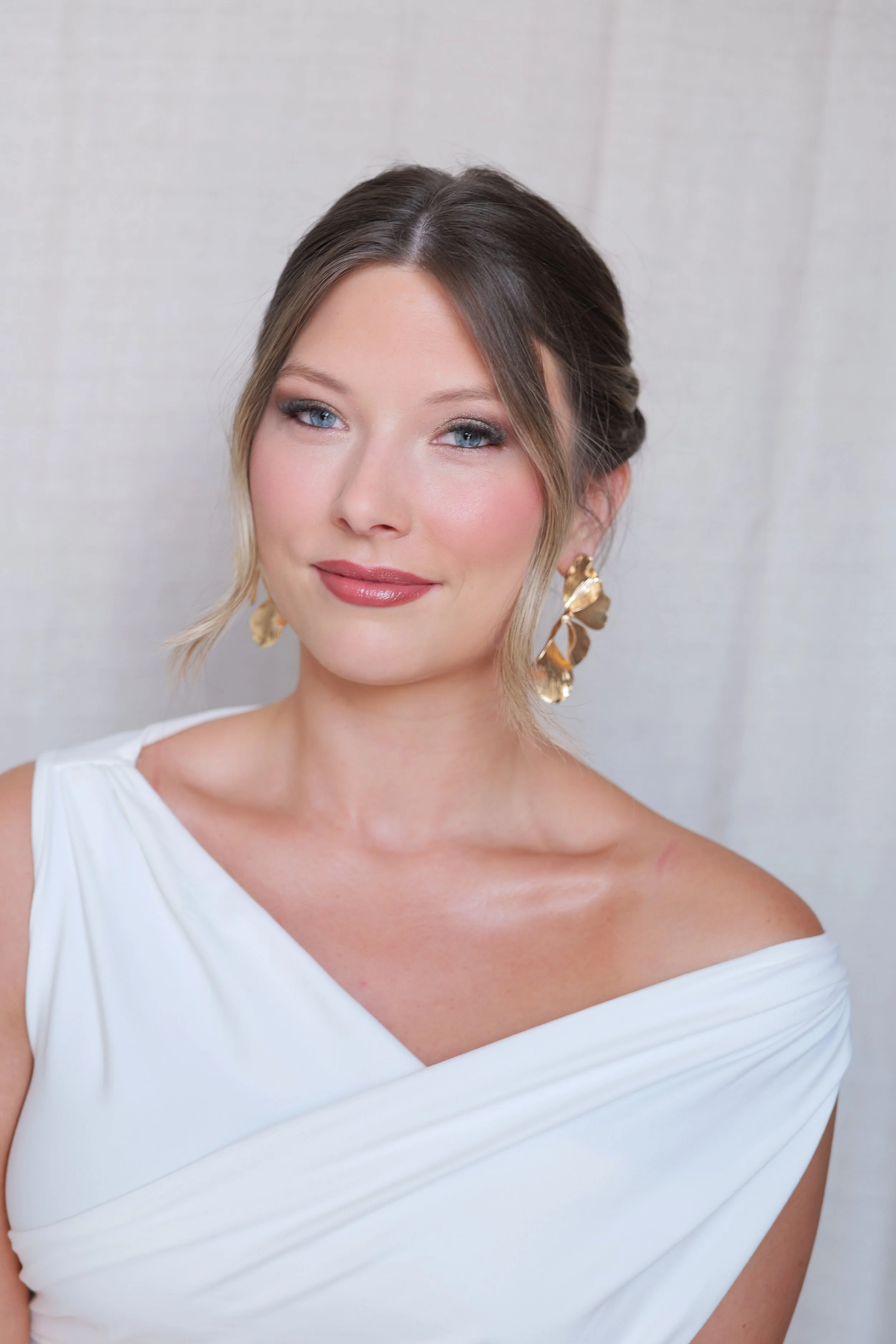 A woman with blue eyes and brown hair styled in an elegant updo, wearing a white off-the-shoulder dress and gold floral earrings, standing against a light background.