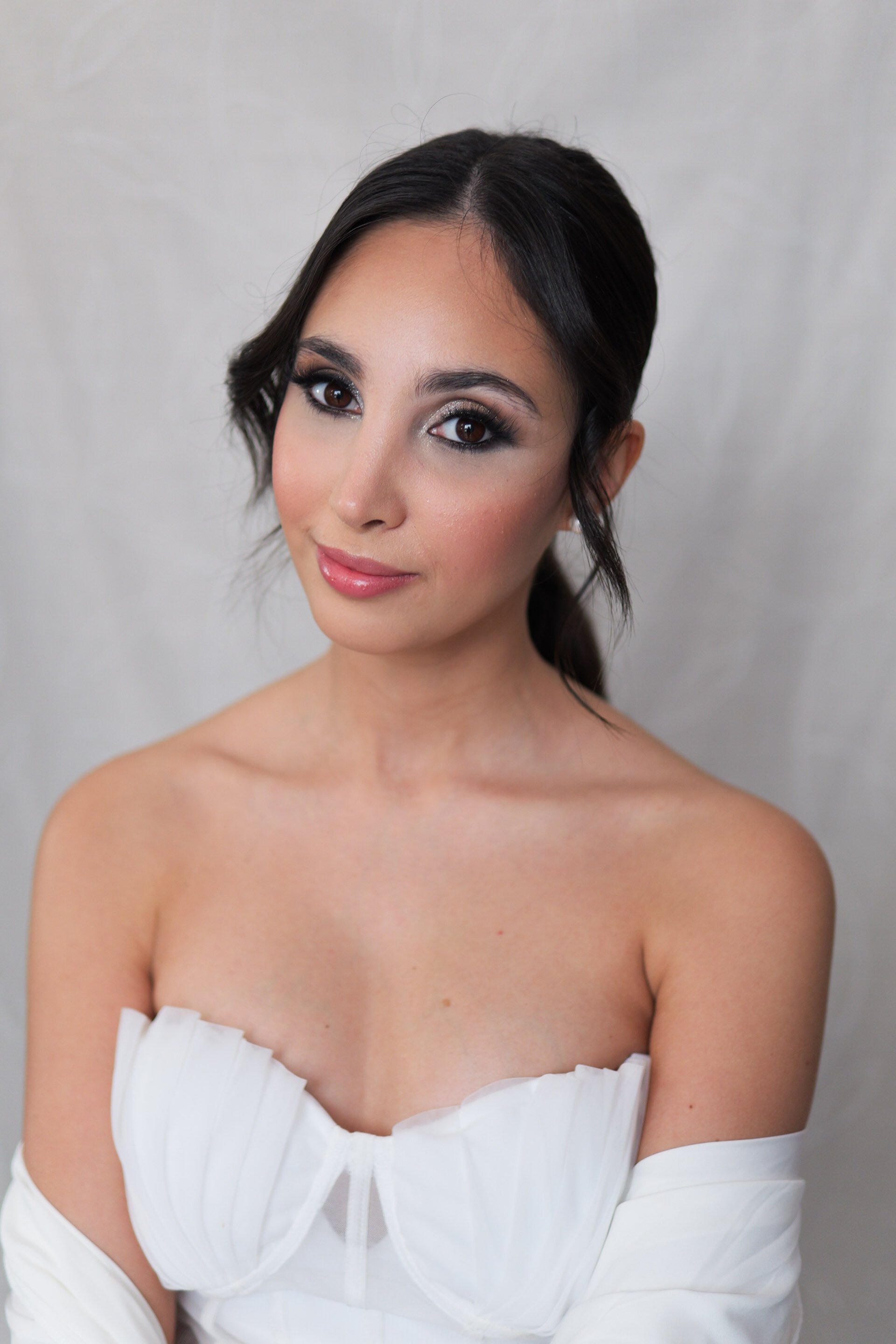 A young woman with dark hair and bridal makeup, wearing a white off-shoulder top, smiling slightly.