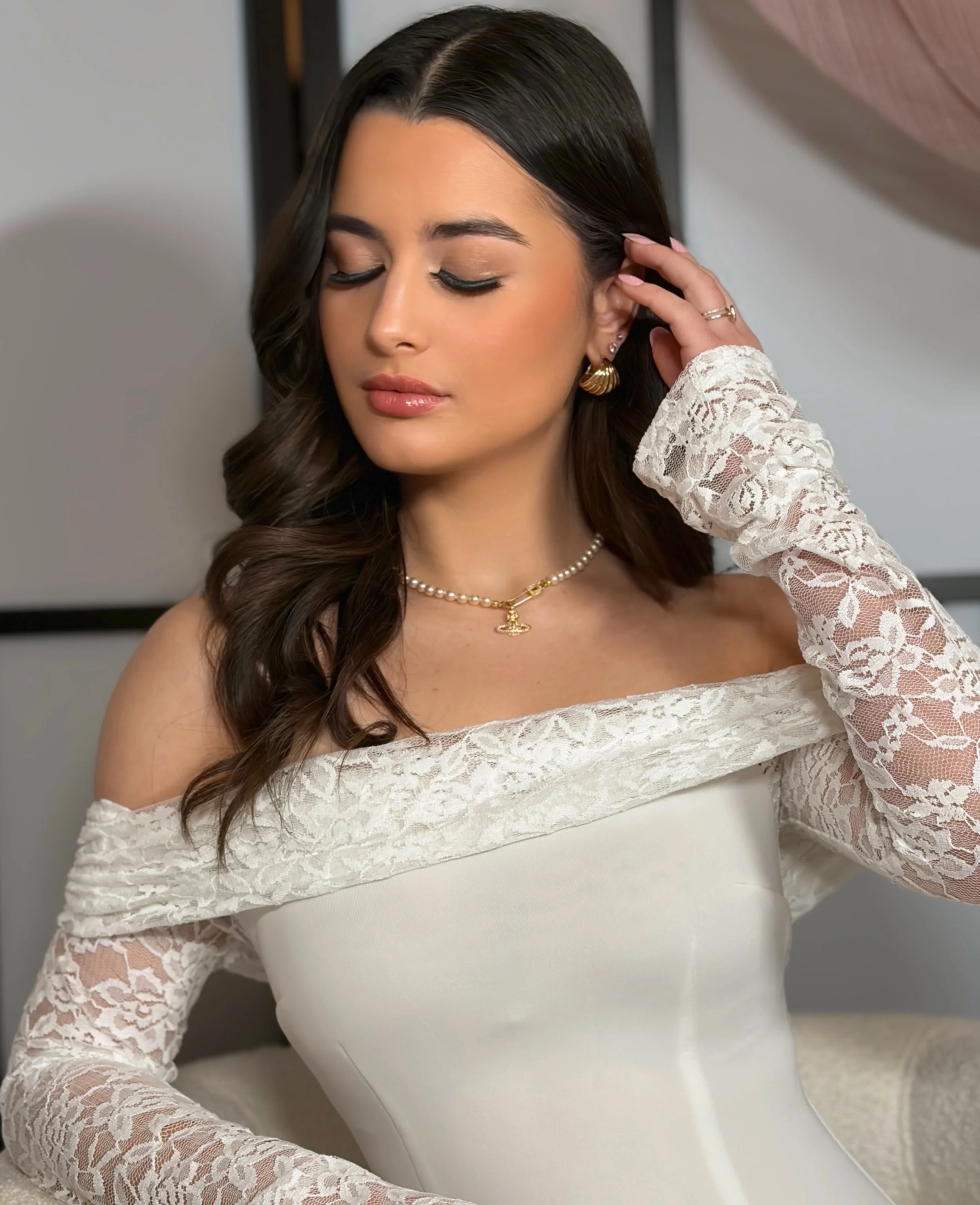 A woman with dark, wavy hair wearing a white lace off-shoulder top and gold jewelry, posing with eyes closed against a neutral background.