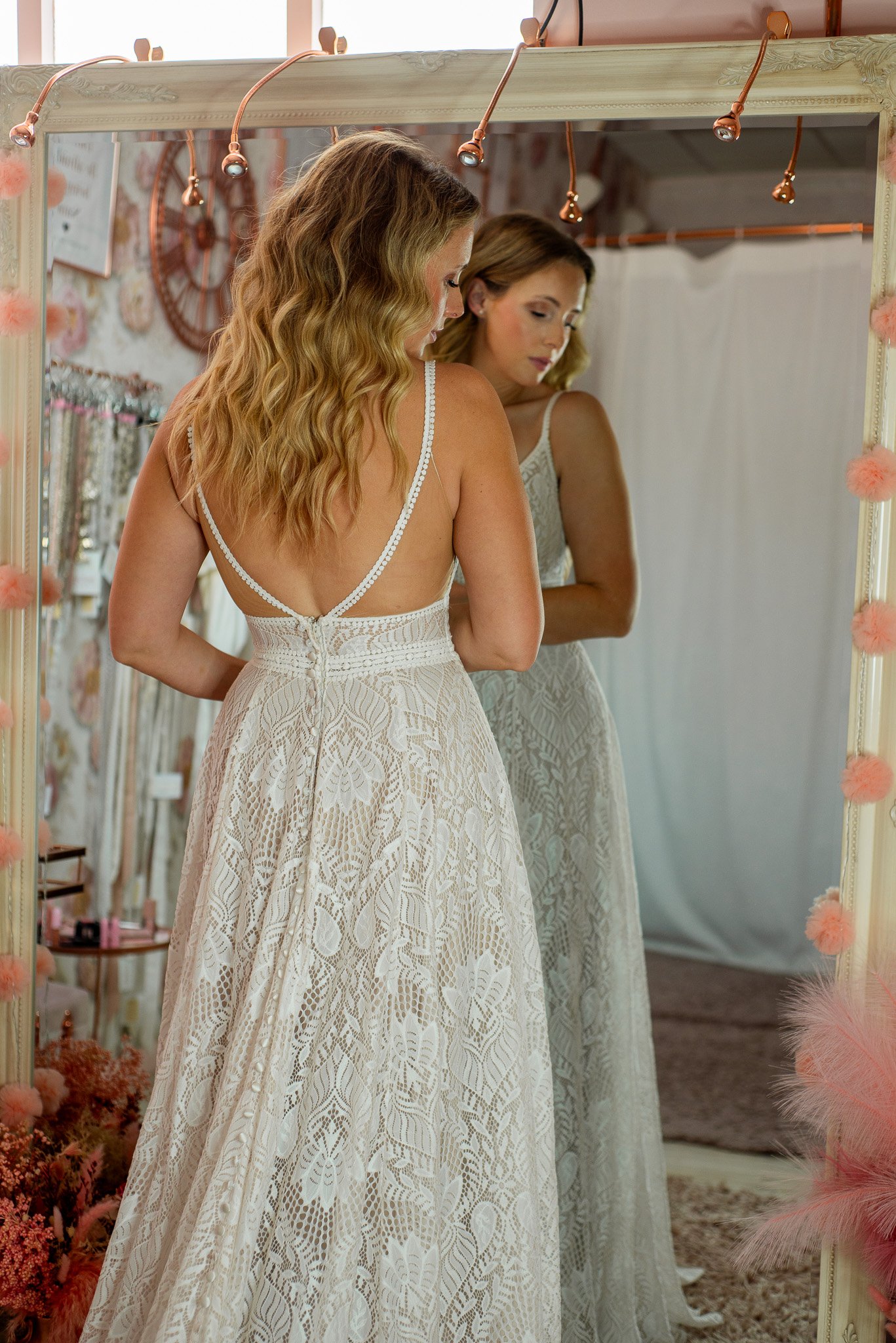 A woman with long wavy blonde hair trying on a cream-colored lace wedding dress with thin straps, looking at her reflection in a large mirror decorated with pink pompoms and soft lighting in a bridal shop.