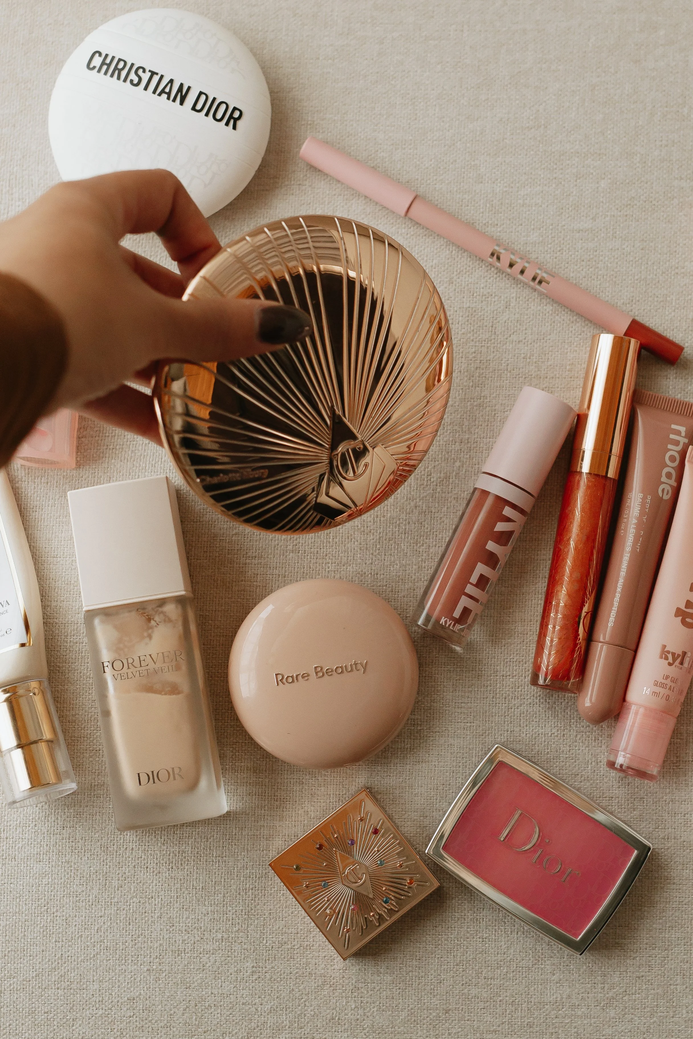 A flat lay of various beauty and makeup products on a beige surface, with a hand holding a metallic rose gold mirror.