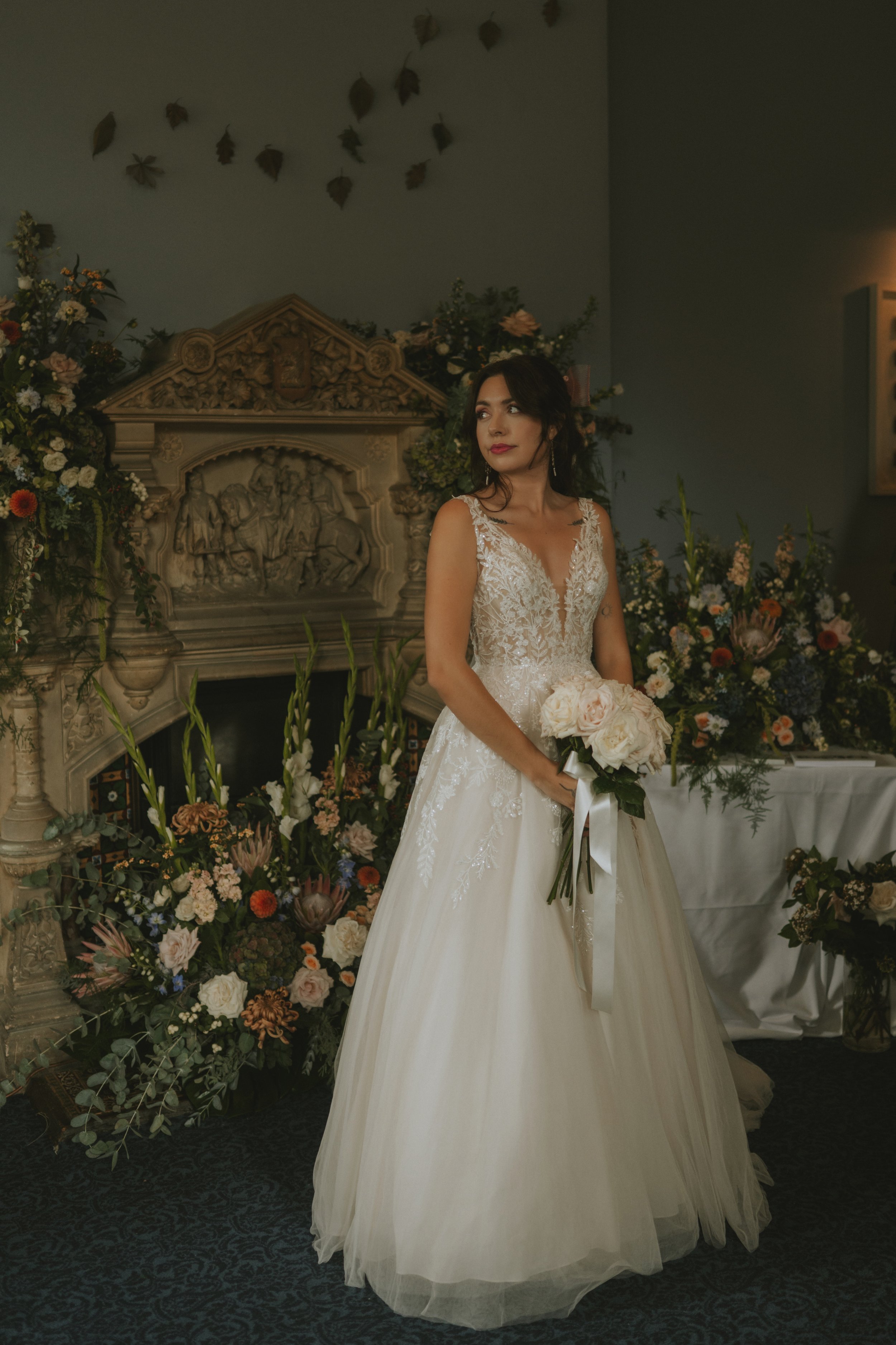 A woman in a wedding dress holding a bouquet of white roses, standing indoors near a decorated fireplace and floral arrangements.