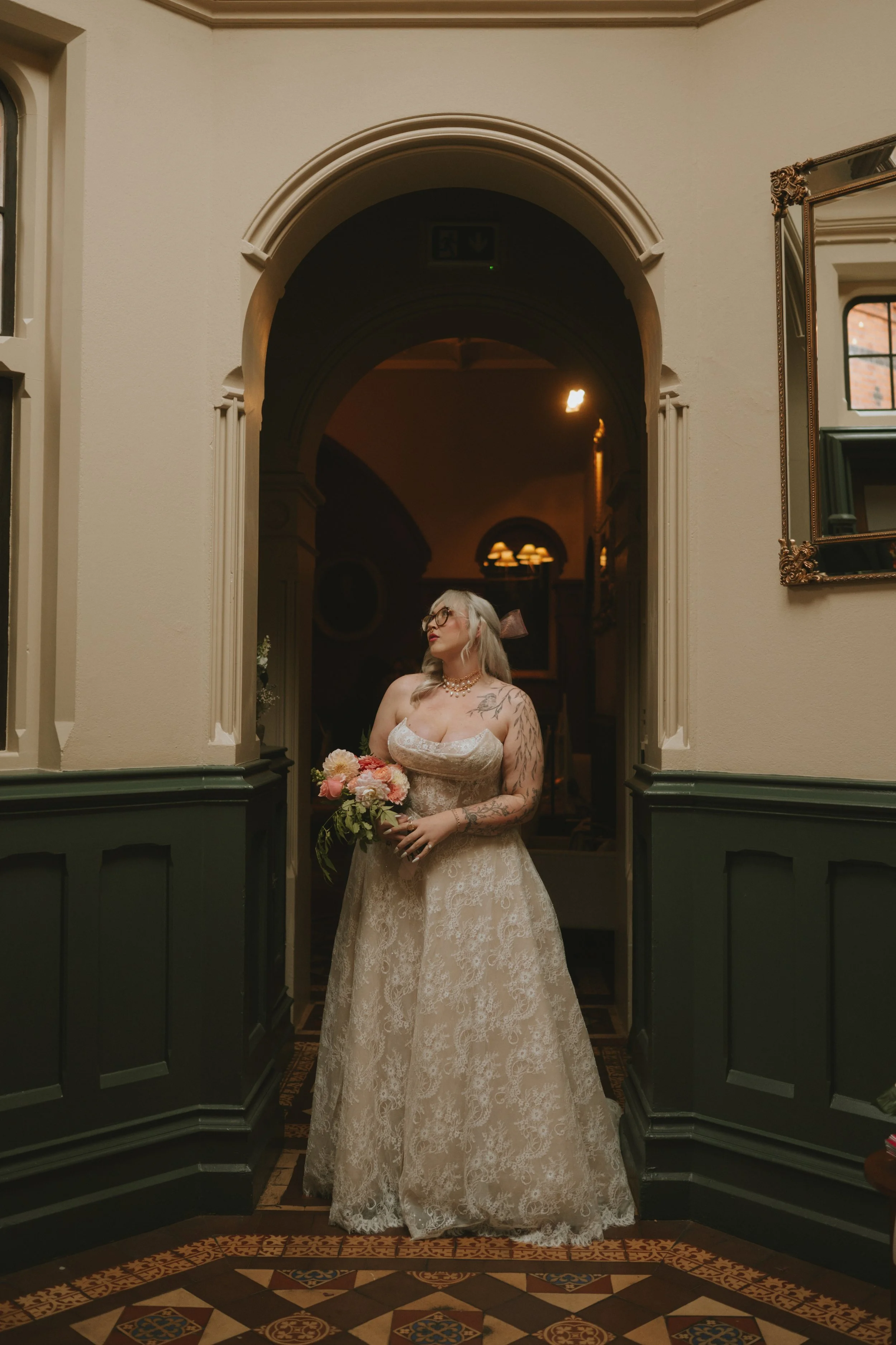 Woman in vintage wedding dress holding a bouquet of flowers, standing in an ornate hallway with dark green wainscoting and patterned tile floor.