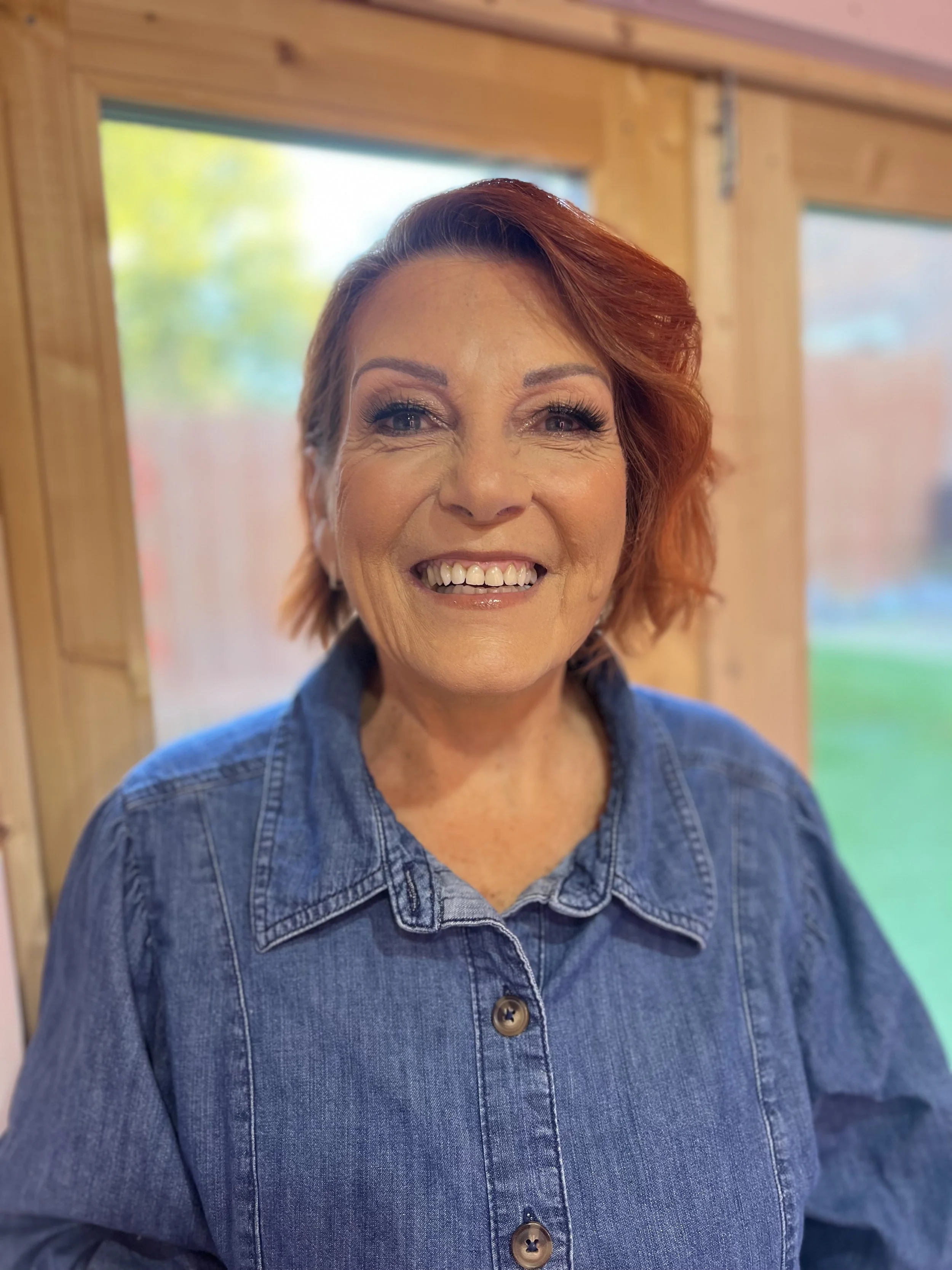Close-up of a smiling woman with red hair, wearing a denim shirt, standing indoors near a window with a blurred outdoor background.