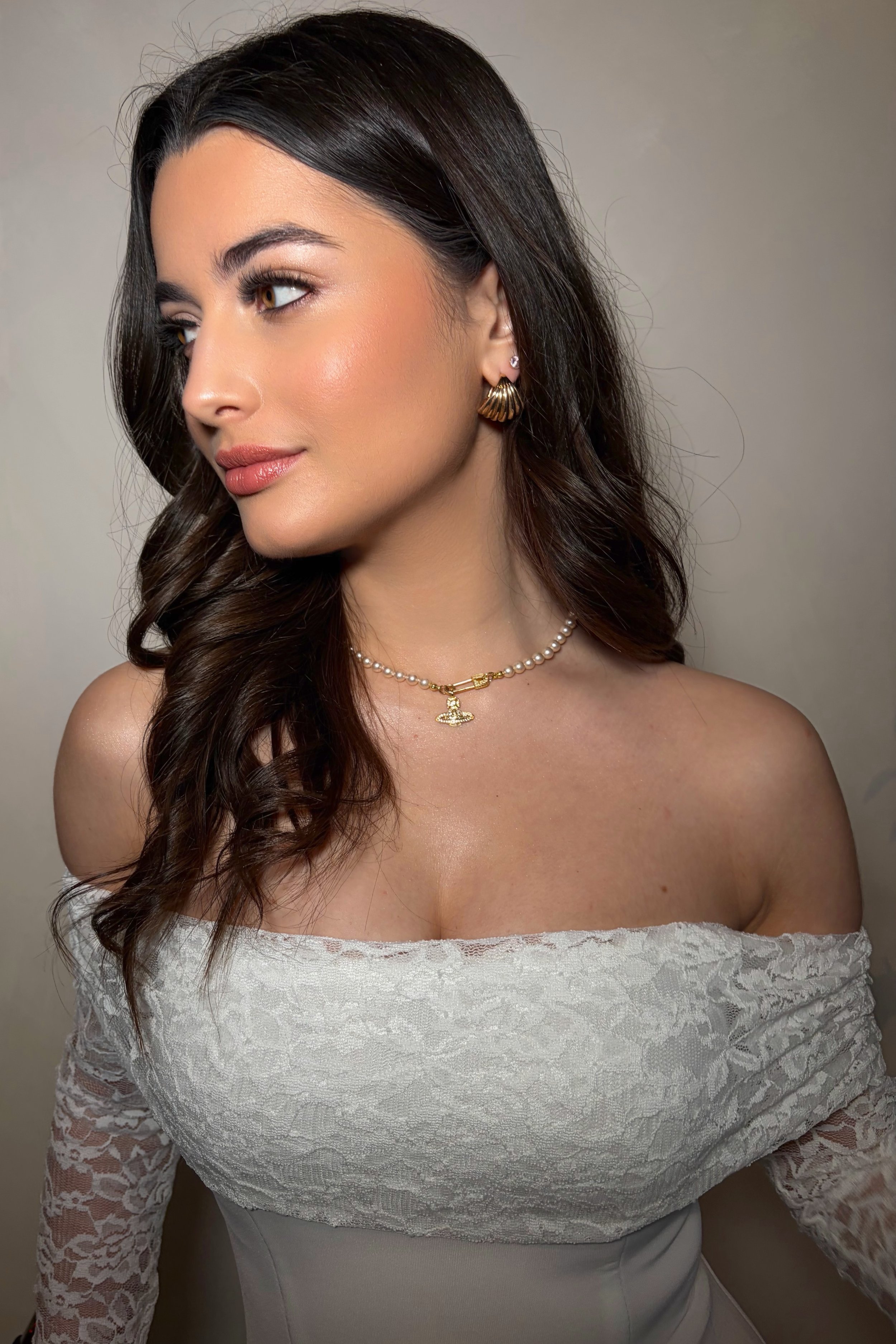 A woman with dark brown hair styled in loose waves, wearing gold hoop earrings, a pearl necklace with a gold pendant, and a white lace off-shoulder dress, standing against a plain background.