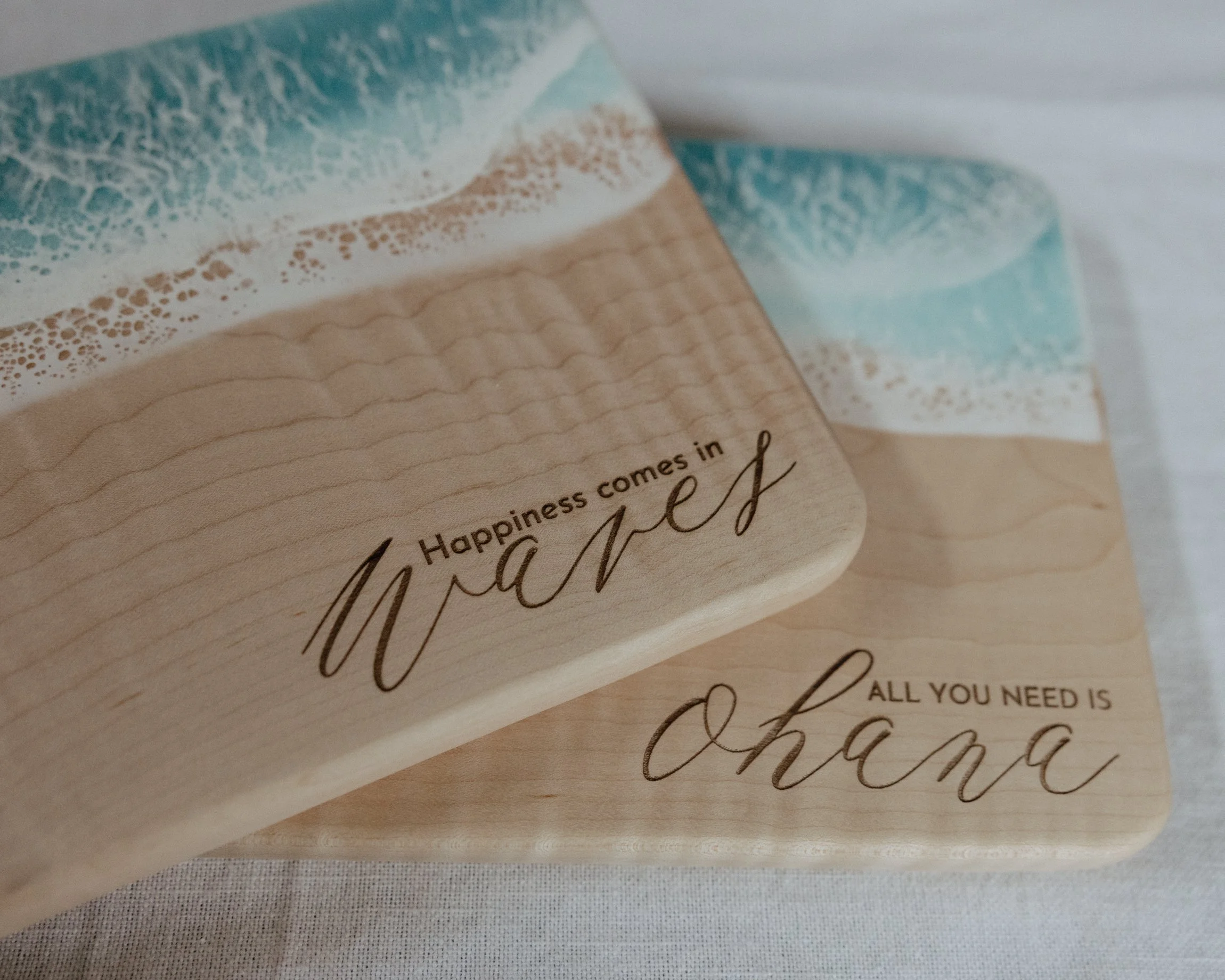Two wooden plaques with ocean wave designs and inspirational quotes, one reads "Happiness comes in waves" and the other "All you need is ohana."