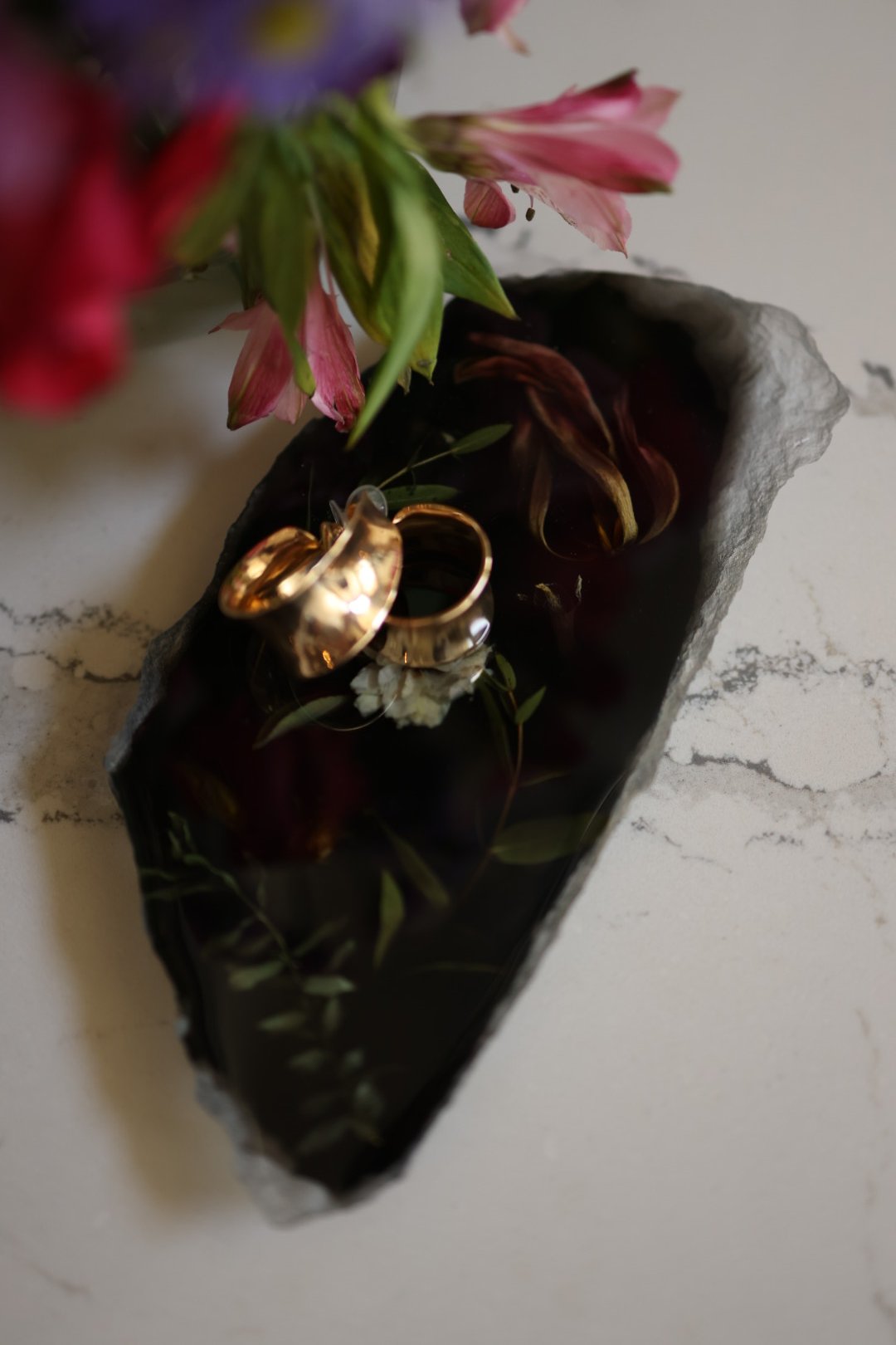Two gold wedding rings placed inside a black decorative tray with scattered white flowers and green leaves, next to a bouquet of pink and purple flowers.