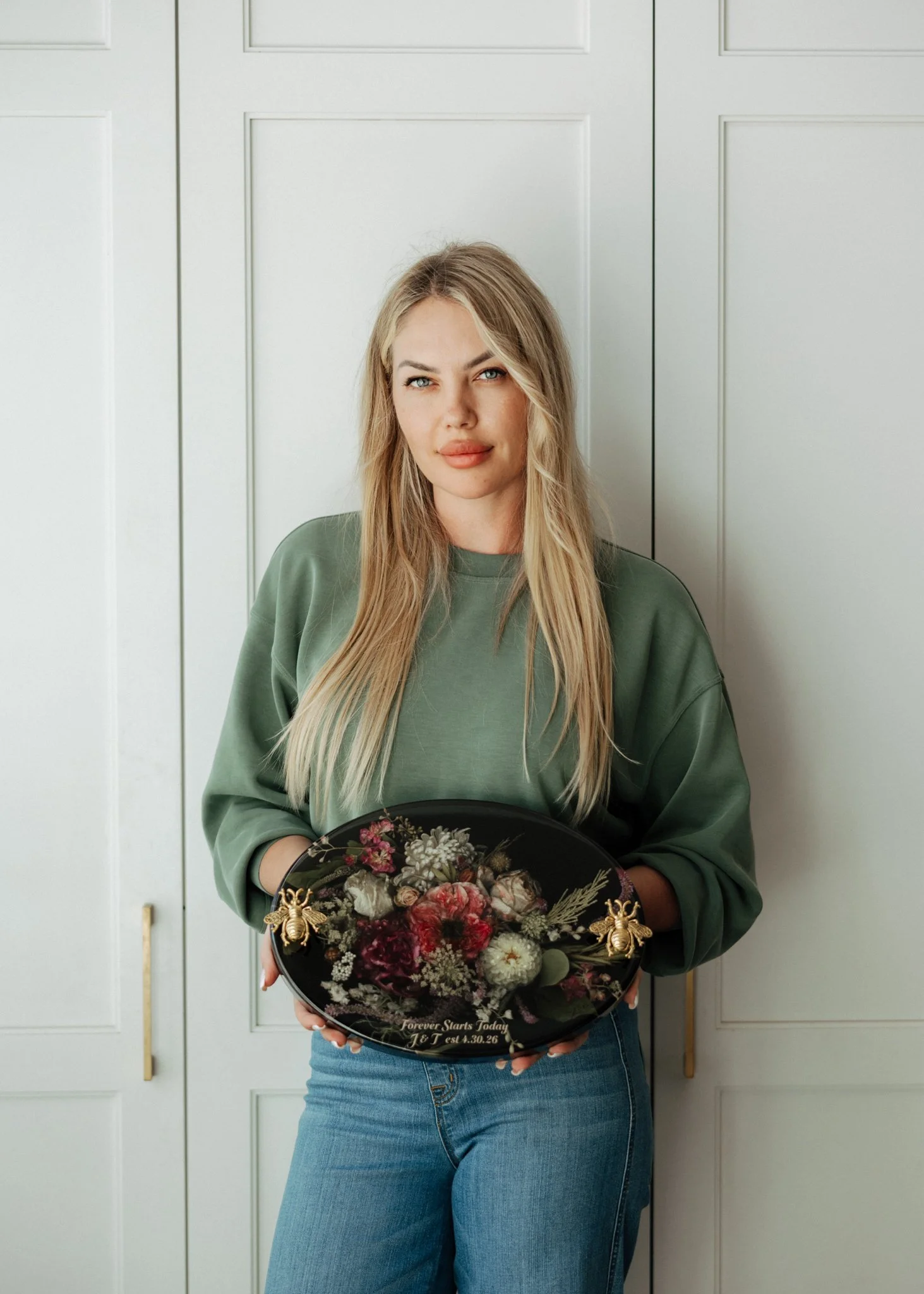 A woman with long blonde hair, wearing a green sweatshirt and jeans, stands in front of white cabinet doors, holding a black tray with flowers and decorative bees, and a message that says "Forever Starts Today" with a date.