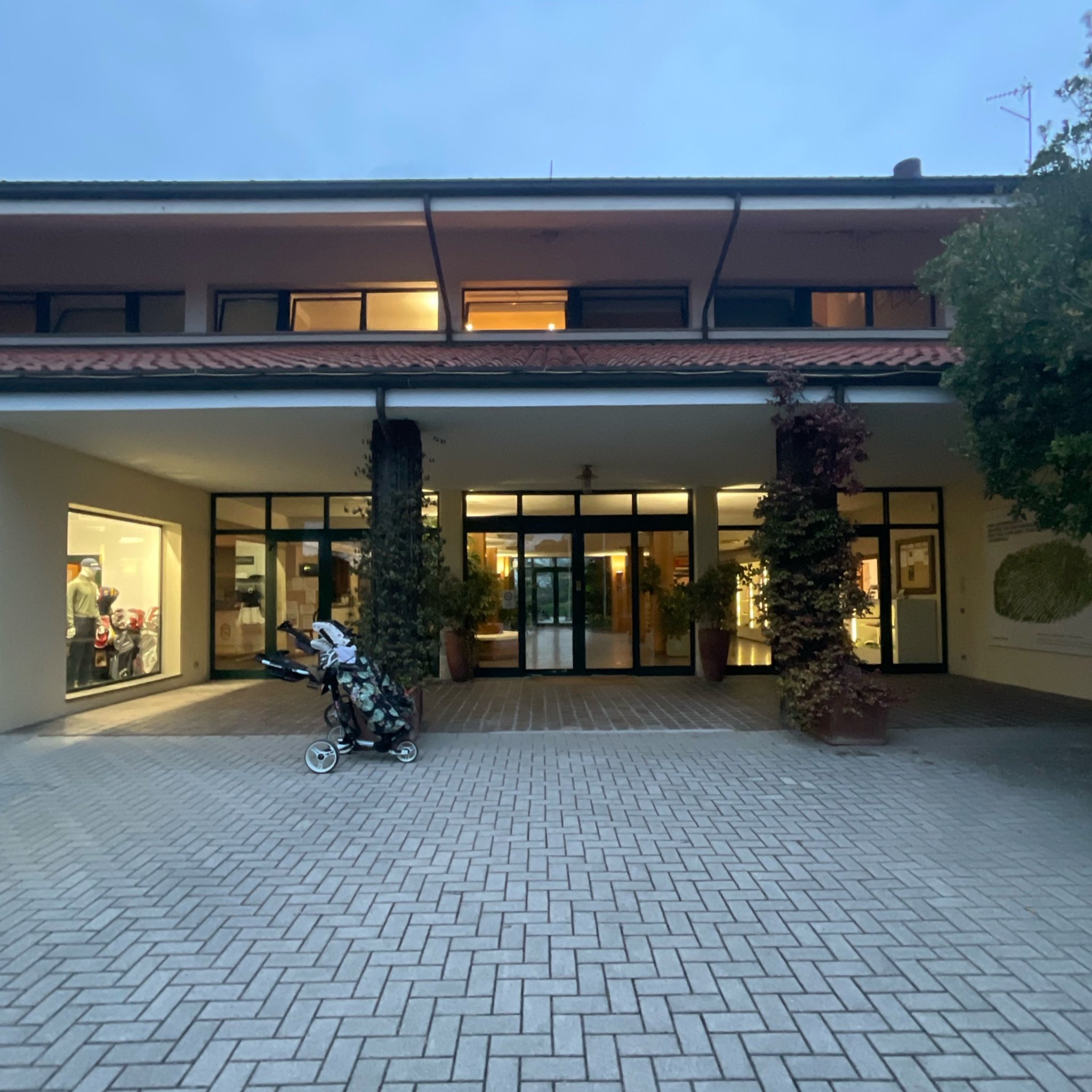 The entrance of a building with glass doors and large windows, a golf bag on a cart in front, potted plants on either side, and parking lot paving in the foreground.