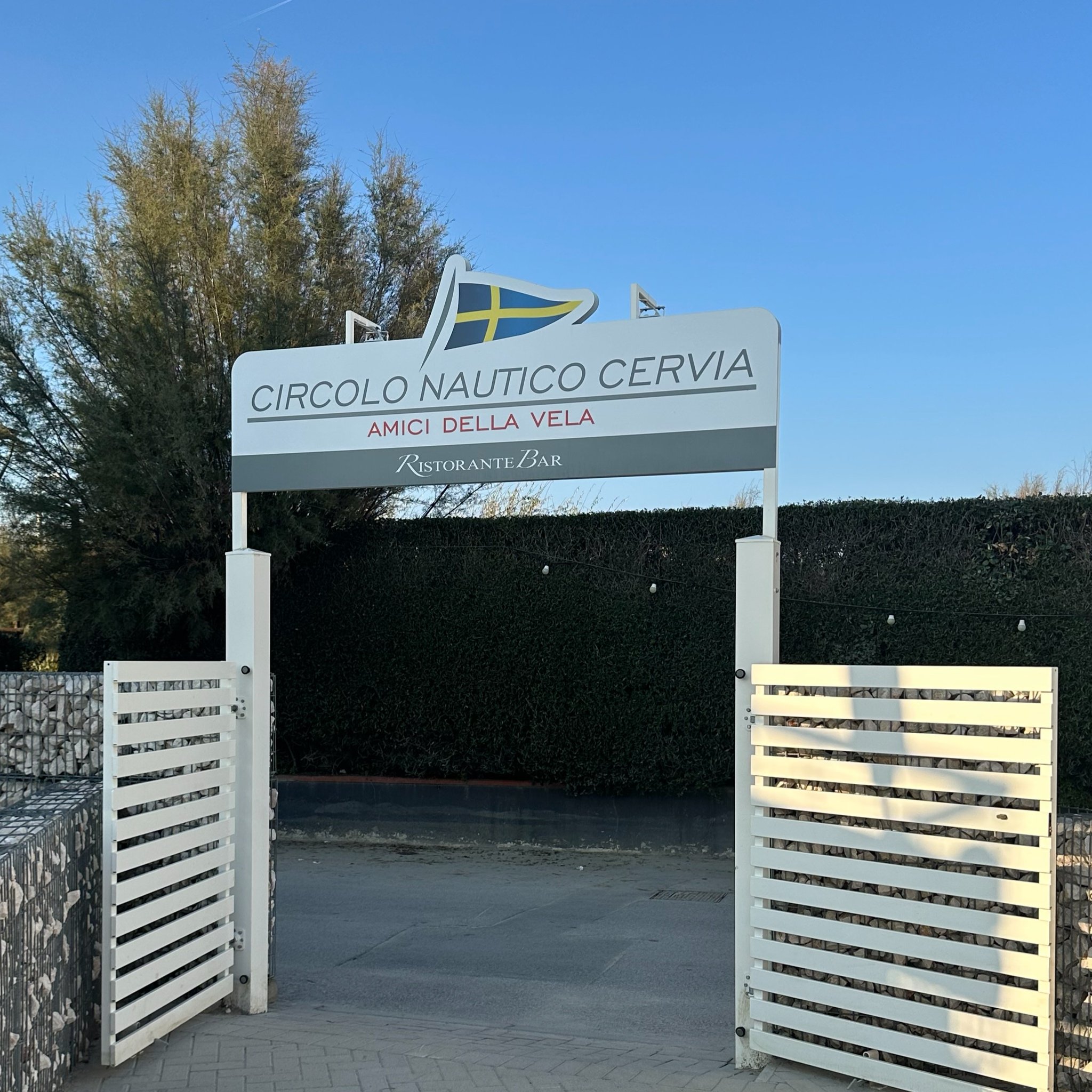 Entrance gate with a sign that reads 'Circolo Nautico Cervia, Amici Della Vela, Ristorante Bar' and a Swedish flag emblem at the top, with trees and a clear blue sky in the background.