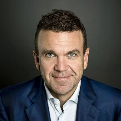 Headshot of a man with short dark hair, blue eyes, wearing a navy suit jacket and white shirt, against a gray background.