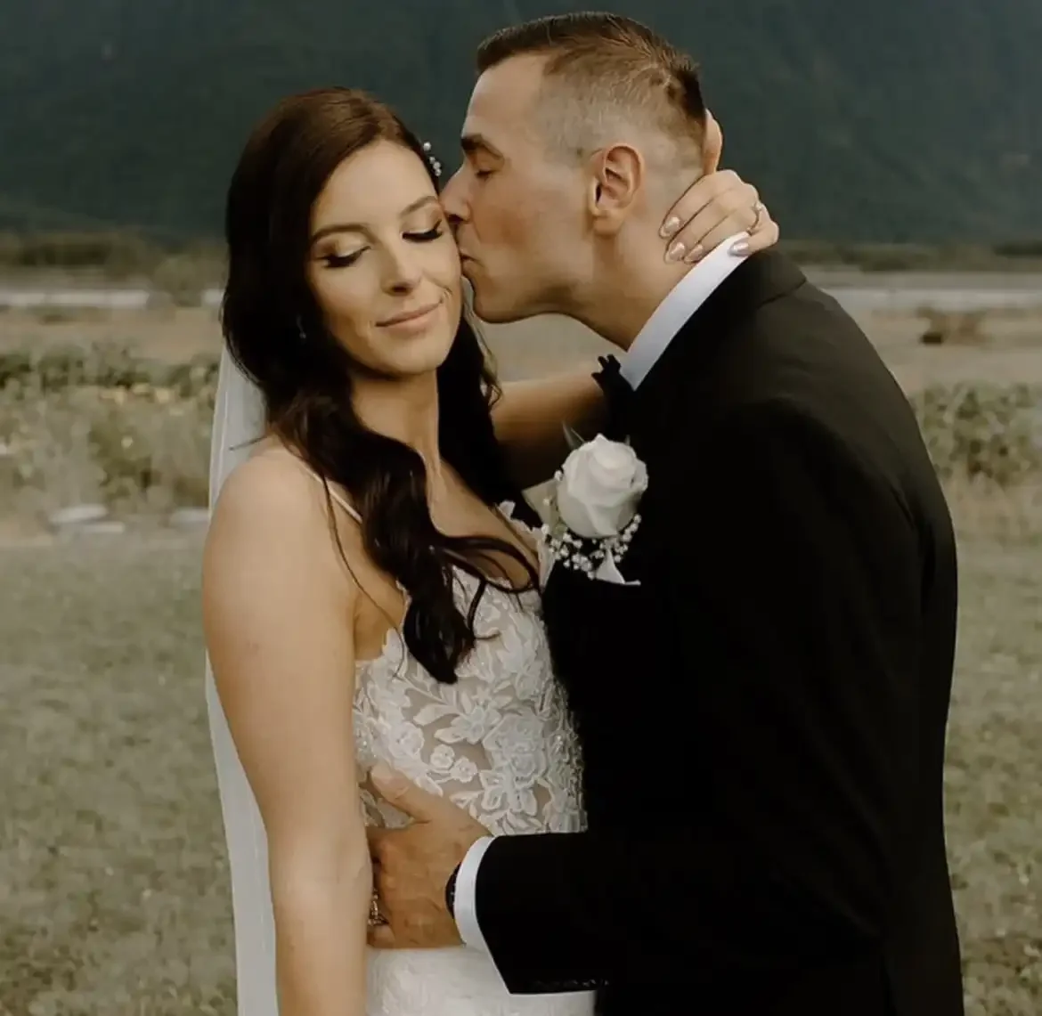 A bride and groom share a kiss outdoors on their wedding day, with the groom gently holding the bride's waist.