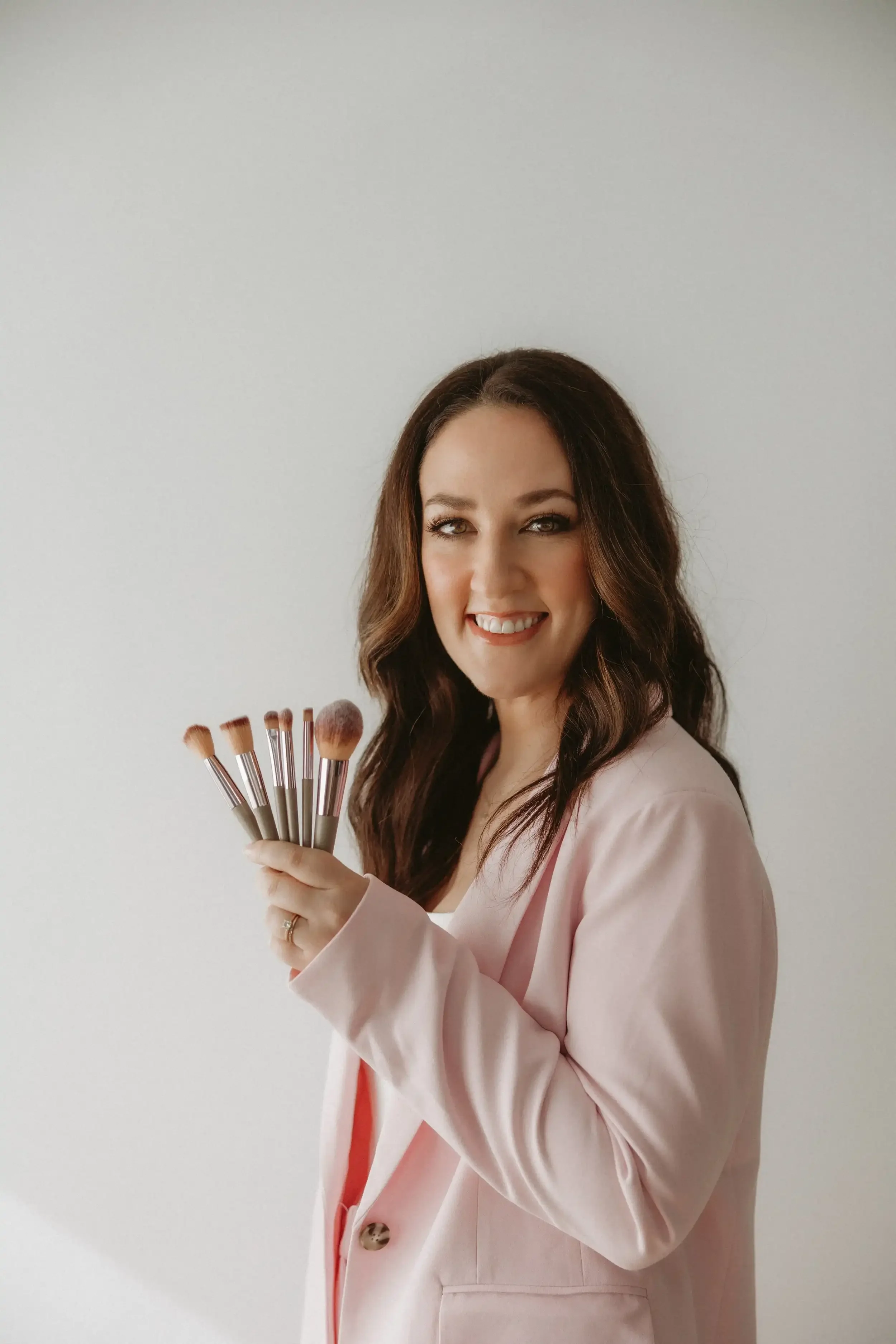 A woman with long brown hair smiling and holding a set of makeup brushes against a plain white background.