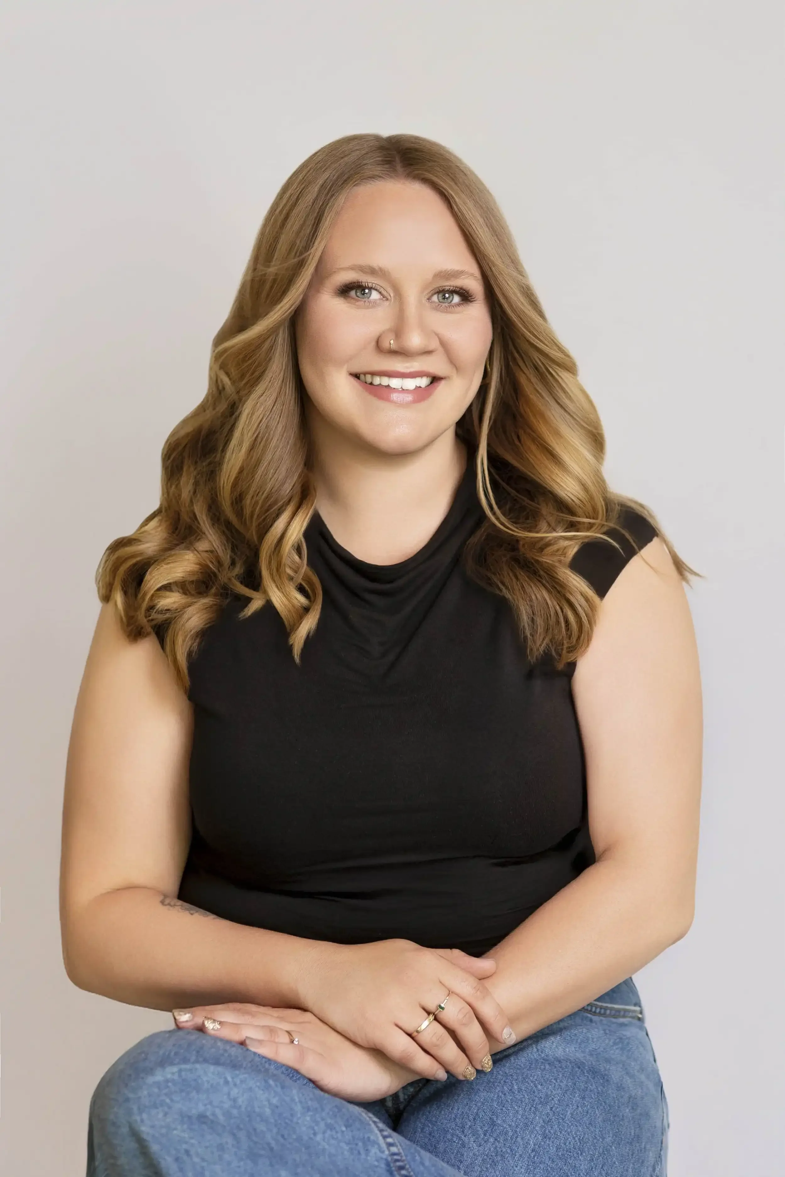 A smiling woman with wavy, light brown hair wearing a black sleeveless top and blue jeans, sitting against a plain light-colored background.