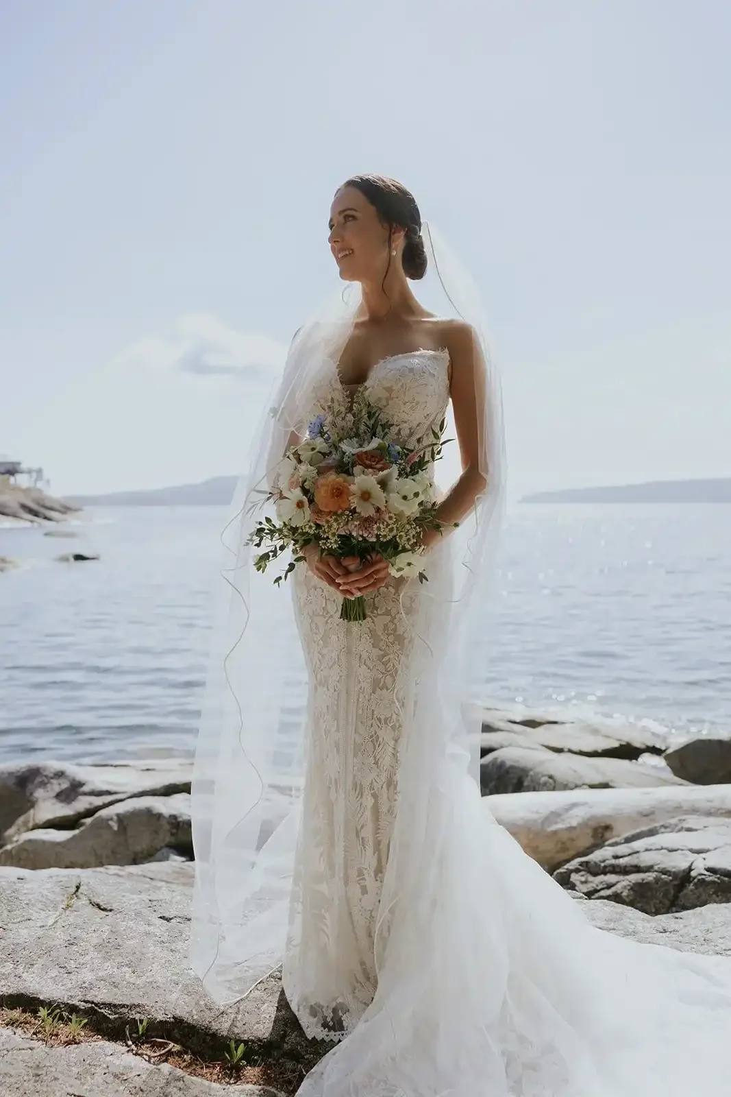 A bride in a lace wedding dress, holding a bouquet, standing on rocks by the water with a scenic ocean view and a partly cloudy sky.