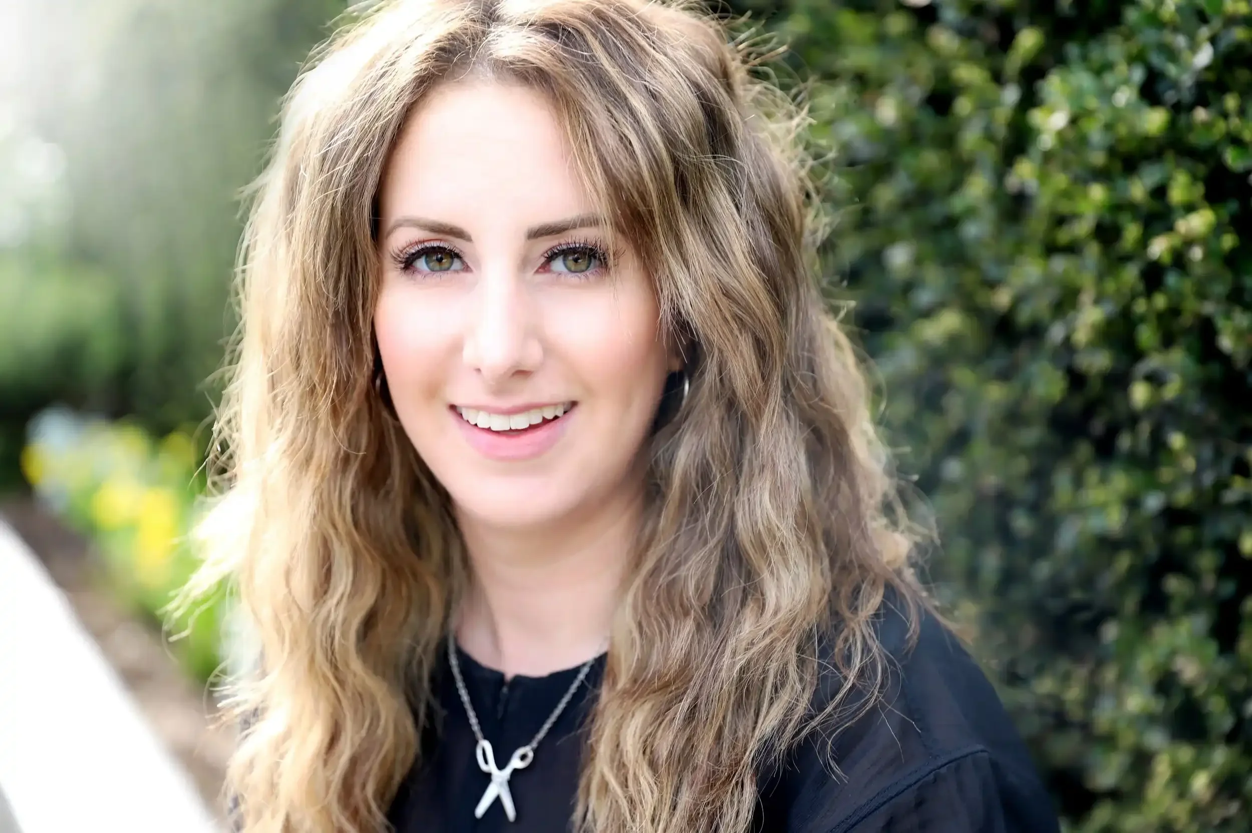 A young woman with long, wavy blonde hair and green eyes smiling outdoors with a green leafy background.