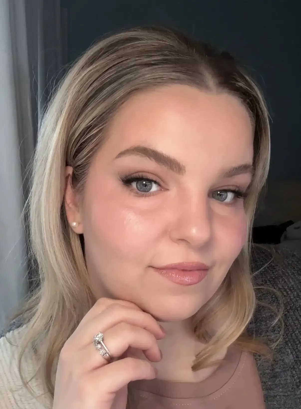Close-up of a woman with blonde hair, blue eyes, and jewelry, smiling softly with her hand resting near her chin.
