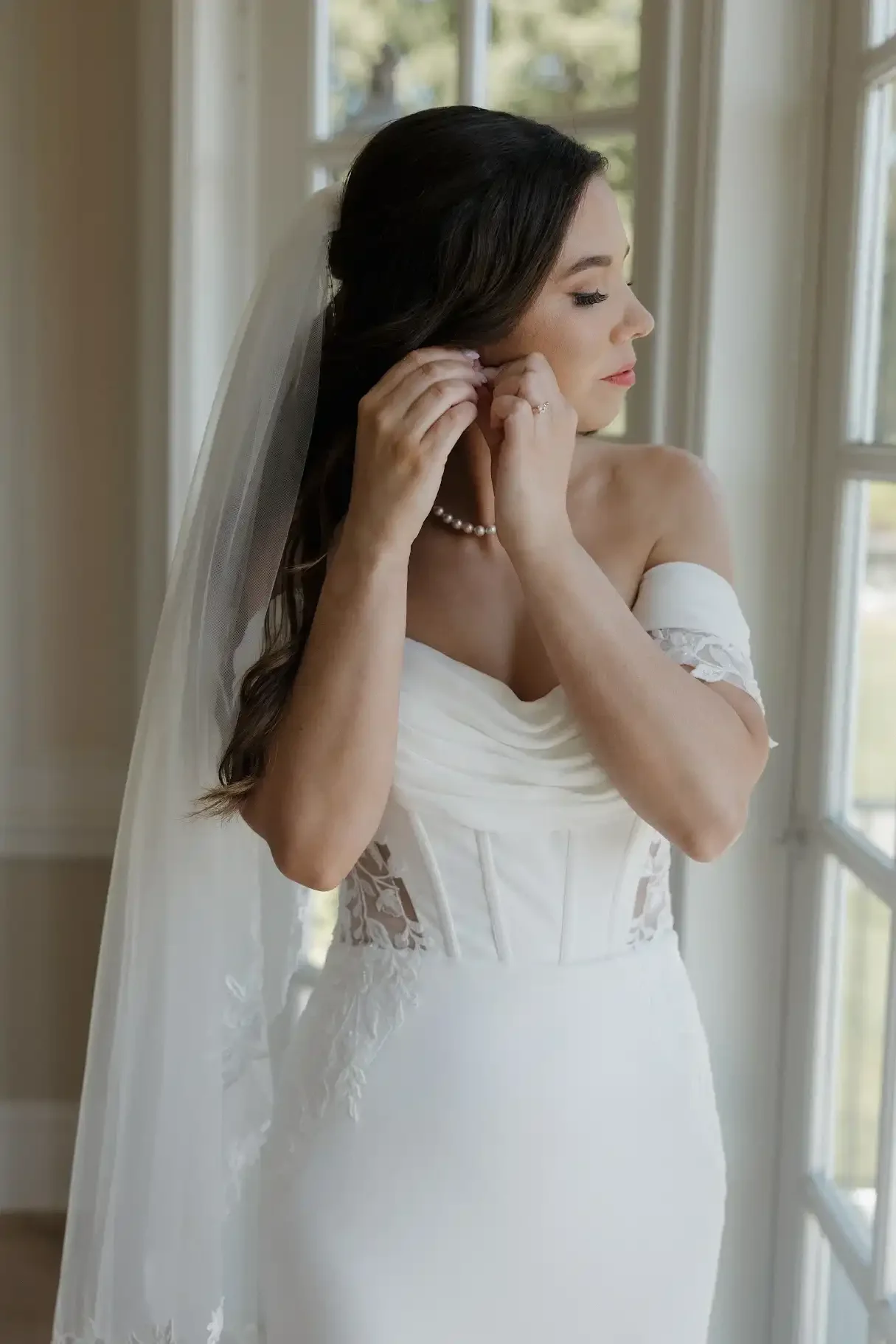 A bride adjusting her earring while standing near a window in her wedding dress.