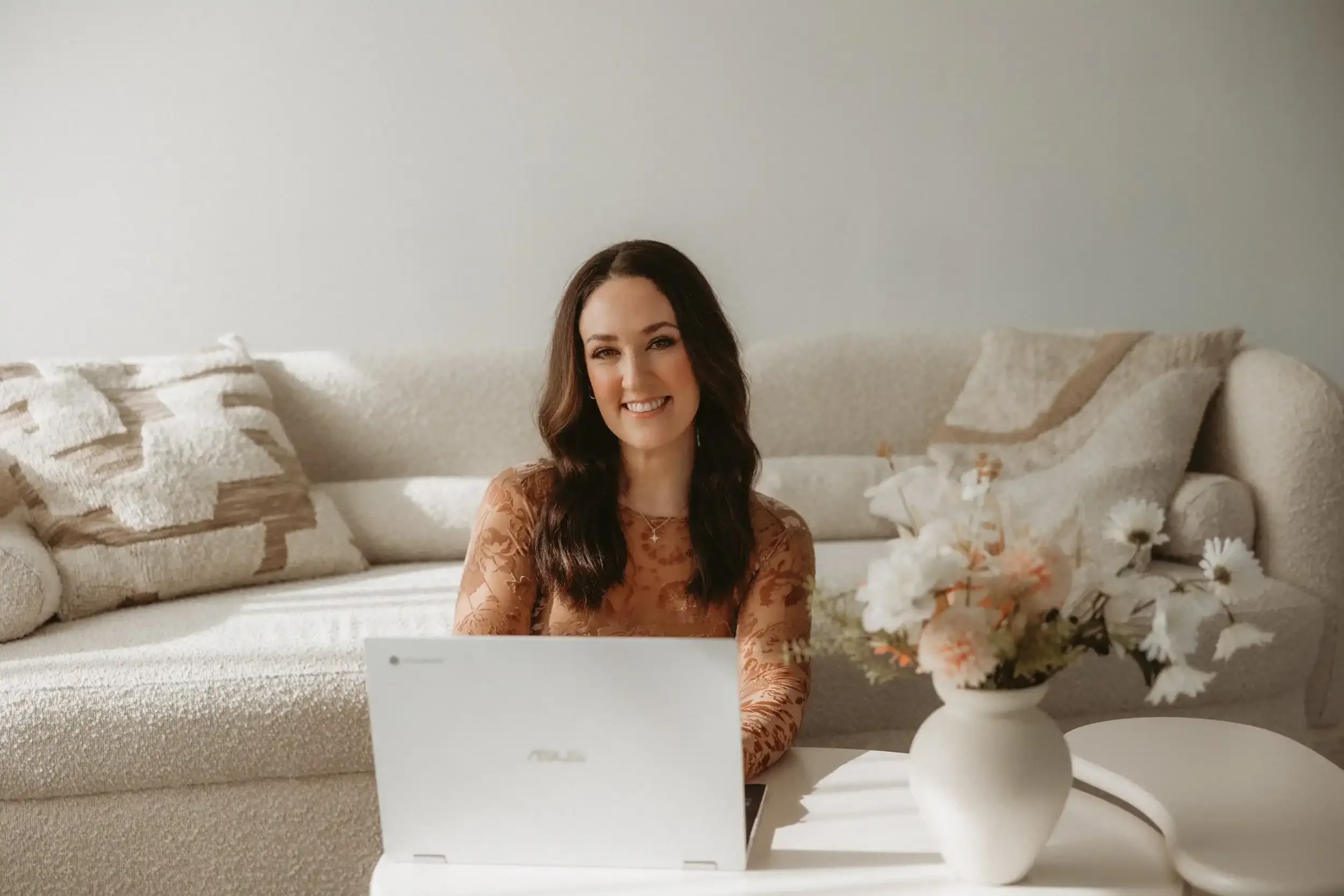 A woman with long dark hair, smiling and sitting on the floor in front of a white coffee table with a white laptop and a white vase with flowers, in a cozy room with white textured sofa and pillows.