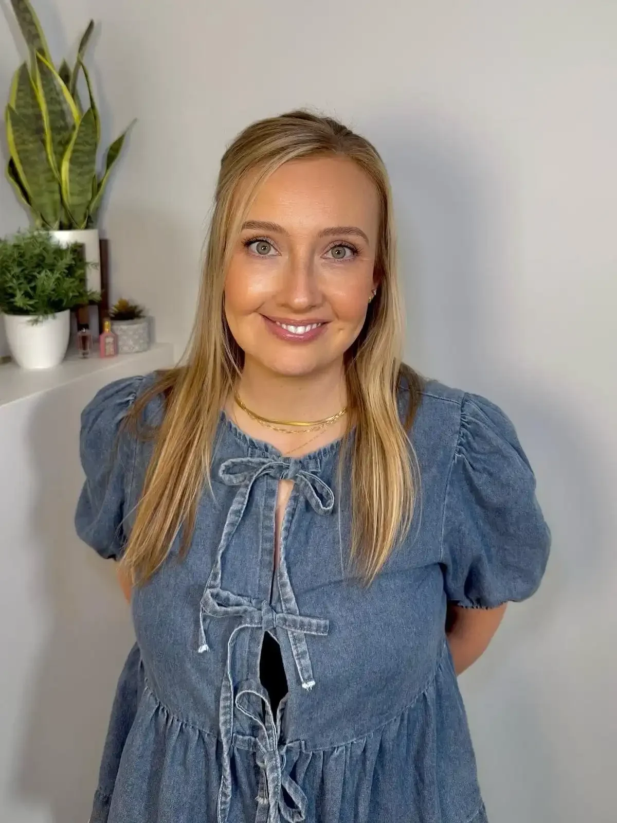 A smiling woman with blonde hair wearing a blue dress with puffy sleeves and a bow tie, standing in front of a plain wall and a shelf with plants and small decor items.