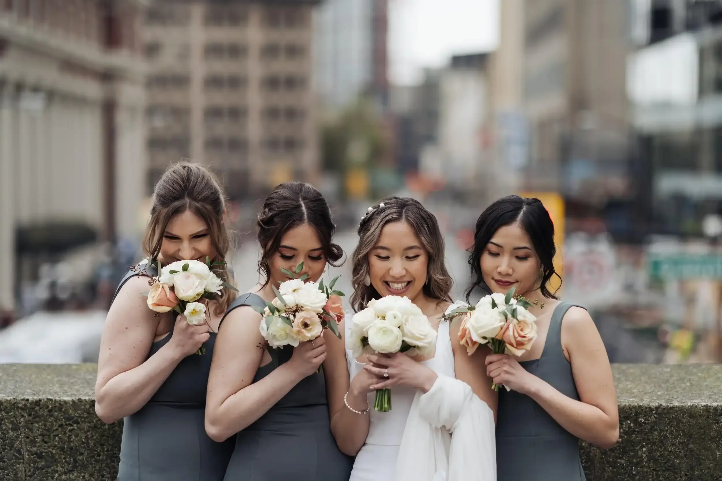 Group of four women in formal dresses holding bouquets of flowers, standing outdoors on a city street.