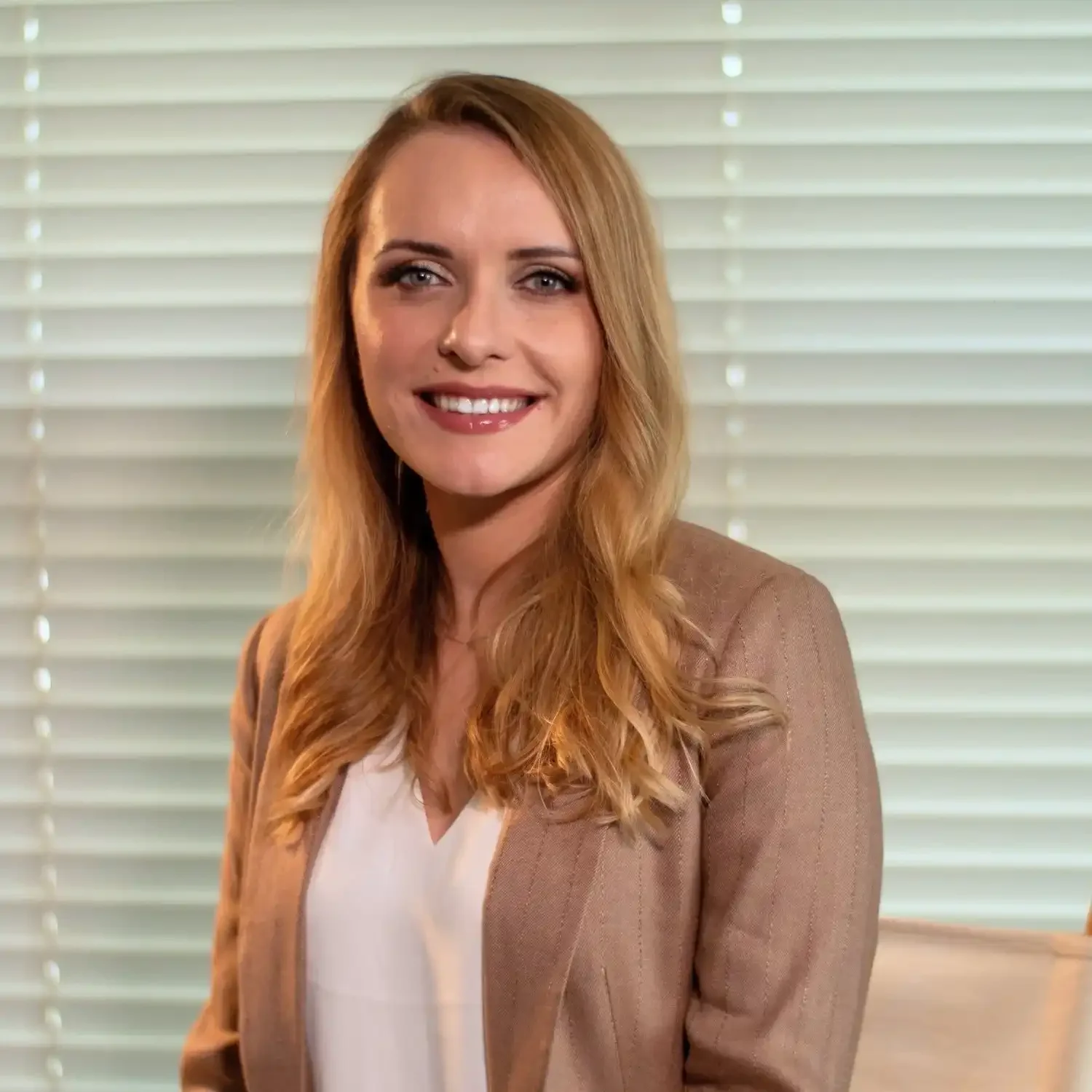 A woman with long, wavy blonde hair, wearing a beige blazer and white top, smiling, with window blinds in the background.