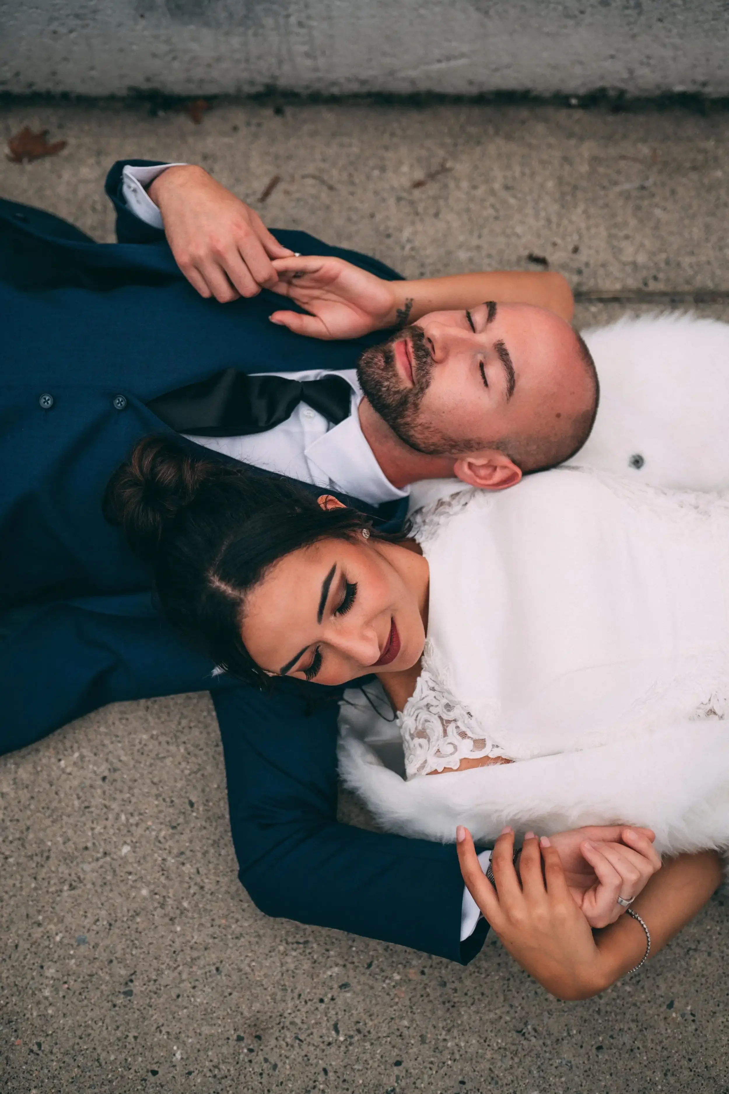 A man and woman lying on the ground, dressed in formal attire; the man is in a suit and tie, and the woman in a white dress with lace details; they appear relaxed, with the woman resting her head on the man's shoulder and their hands intertwined.