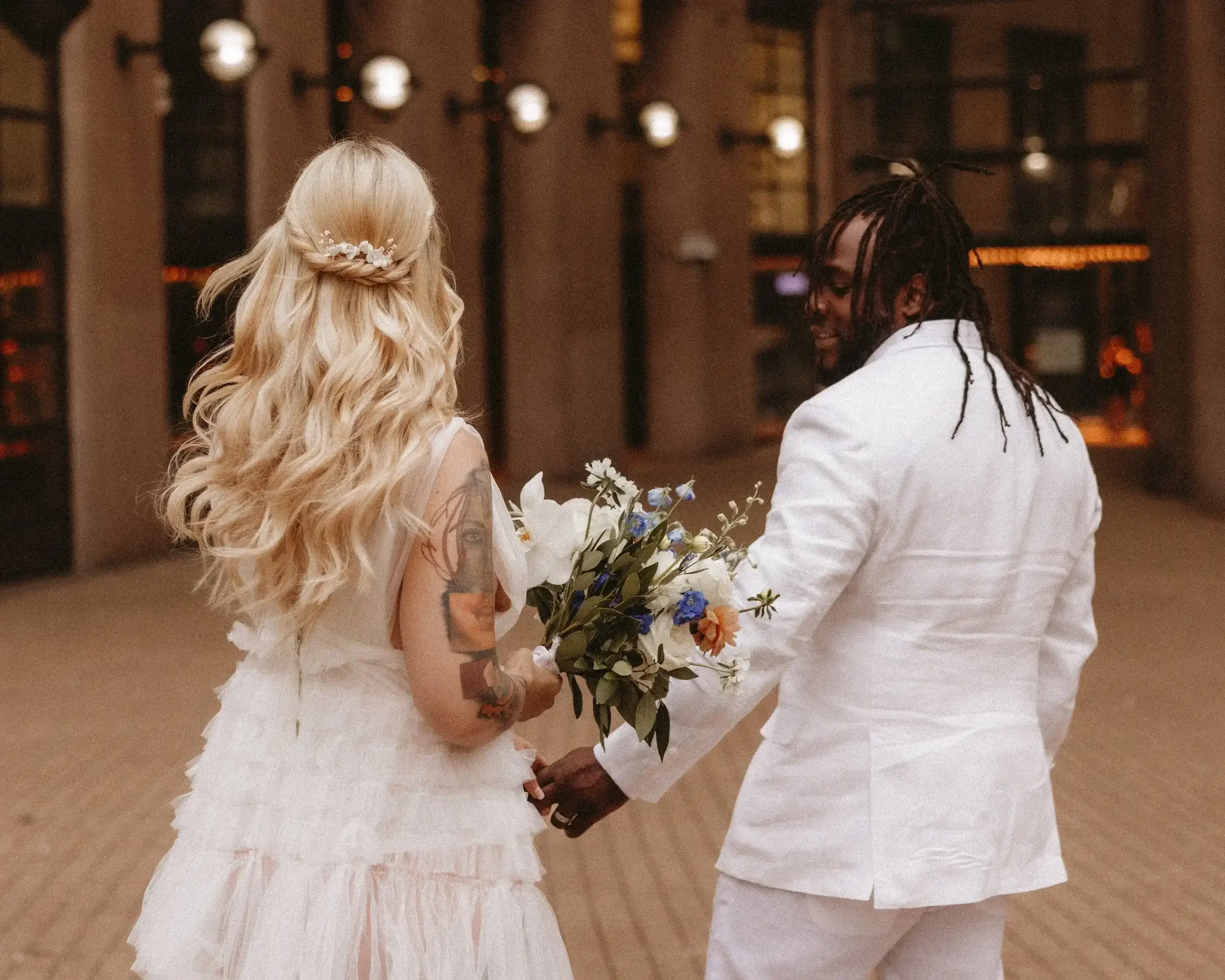 A couple dressed in white wedding attire holding hands and a bouquet, standing in an urban, lit corridor at night.