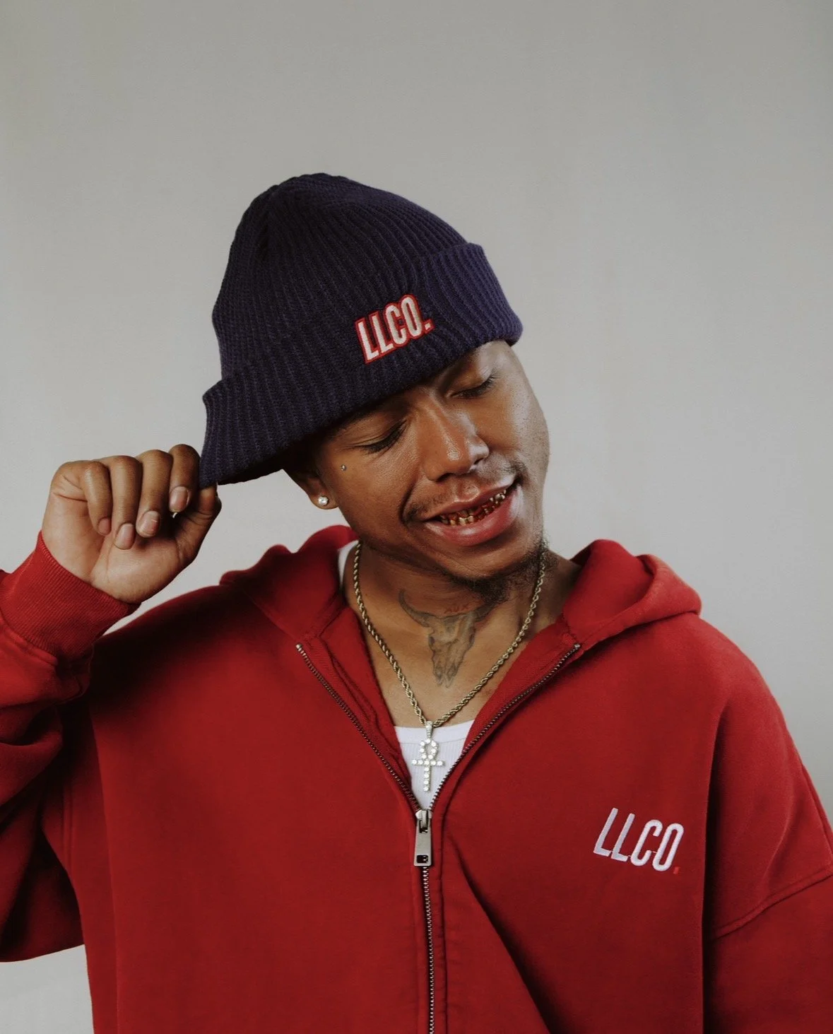 A young man wearing a red hoodie and a navy blue beanie with red and white 'LLCO' logo, touching the brim of his hat. He has gold teeth, earrings, a tattoo on his neck, and is smiling.