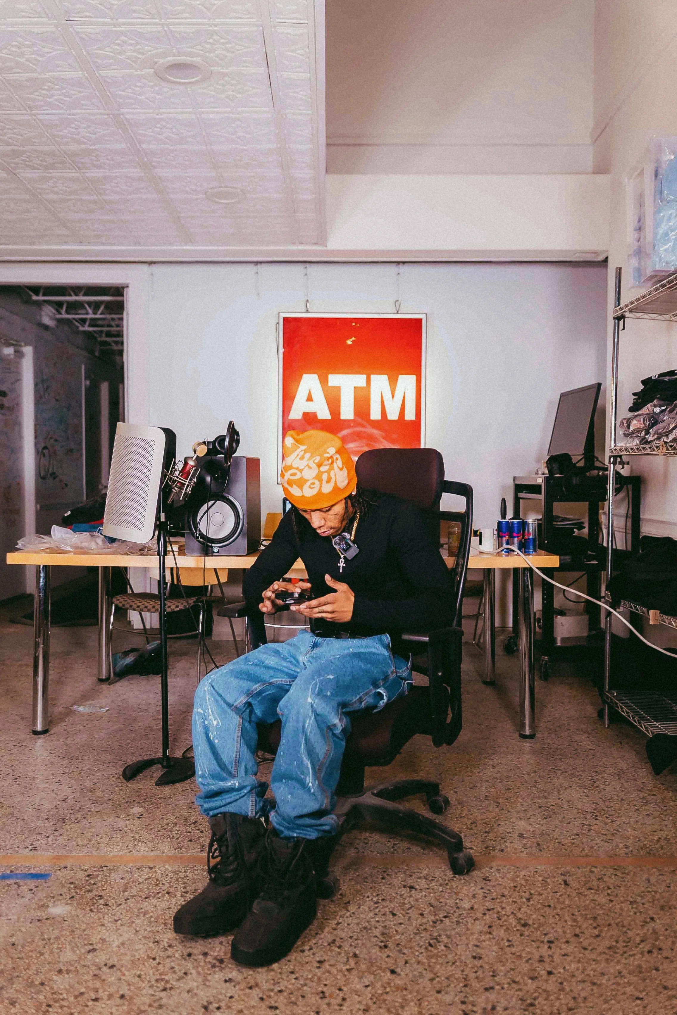 Person sitting in an office chair, looking at a phone, wearing a Orange beanie says Just Love, black shirt, baggy jeans, and black boots. Behind them is a desk with audio equipment, a speaker, and a computer monitor, with an 'ATM' sign on the wall.