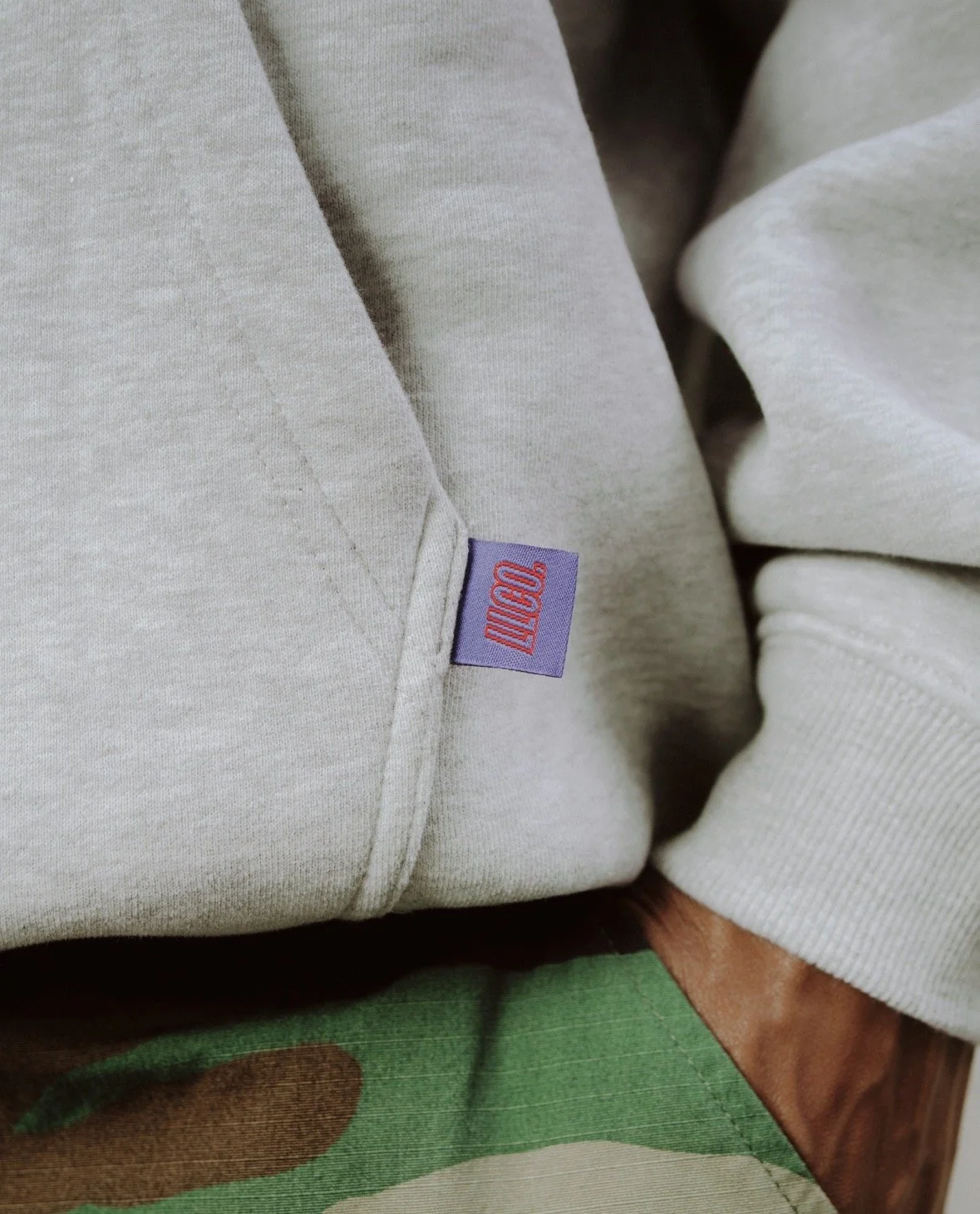 Close-up of a beige hoodie with a purple tag that has red text reading 'LLCO.' near the seam.