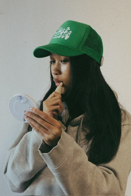 A girl wearing a green cap with white lettering that says Just Love, looking at a handheld mirror and touching her lips with her index finger.