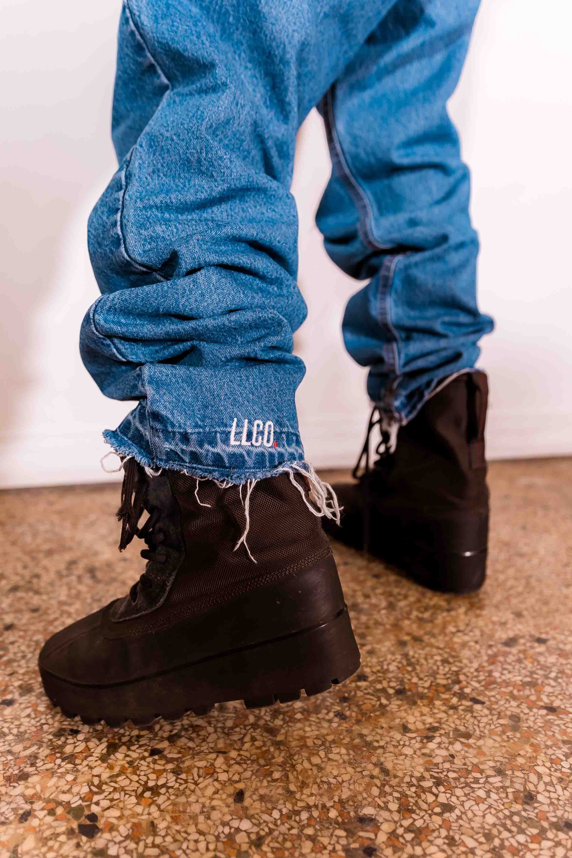 Close-up of a person's legs wearing ripped blue jeans with frayed cuffs with LLCO. embroidered and black winter boots, standing on a speckled floor with a white background.