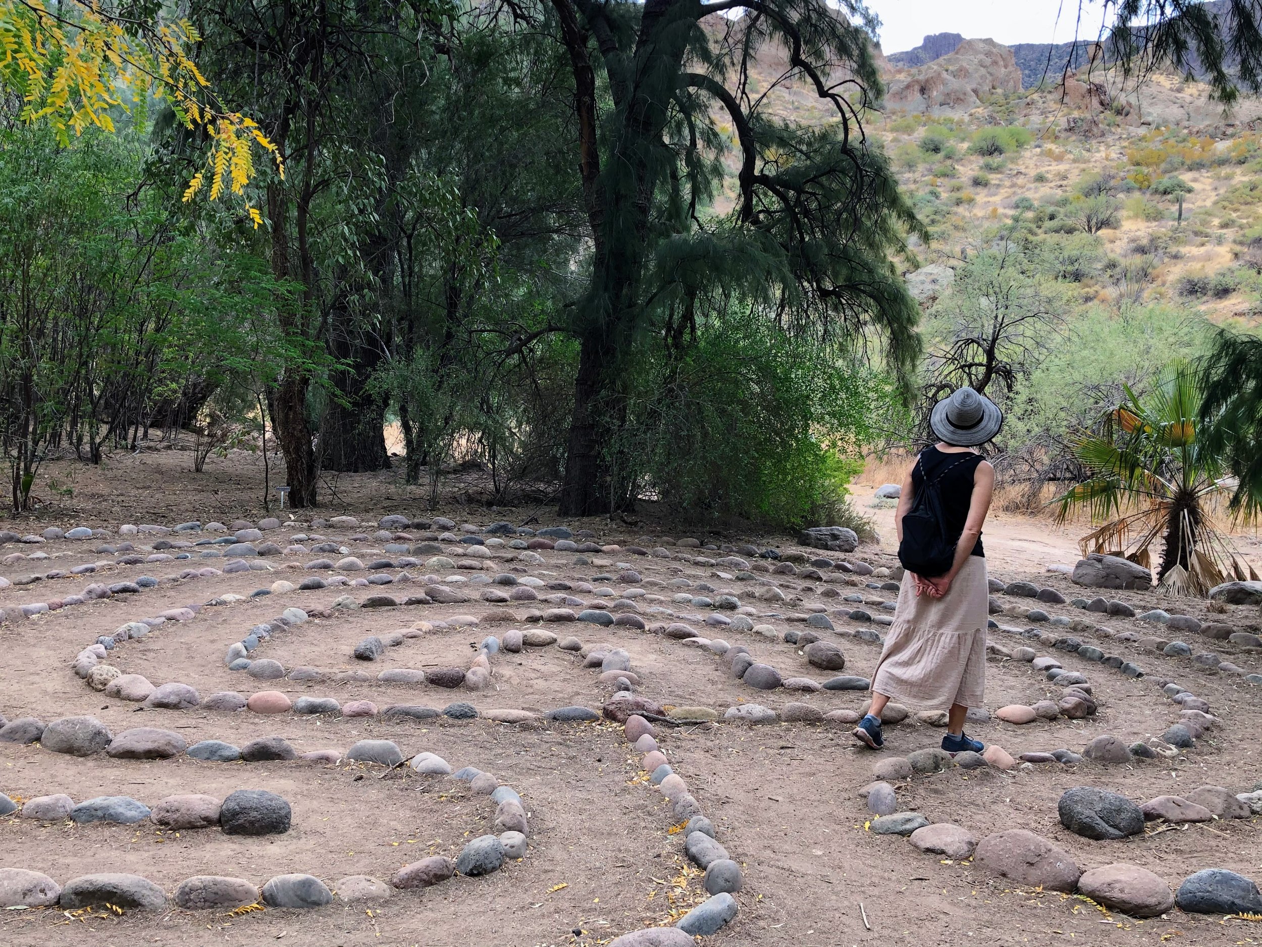 A woman wearing a large sunhat, black top, beige skirt, and blue shoes walks through a circular pattern of rocks on the ground in a desert landscape surrounded by trees, bushes, and distant hills.