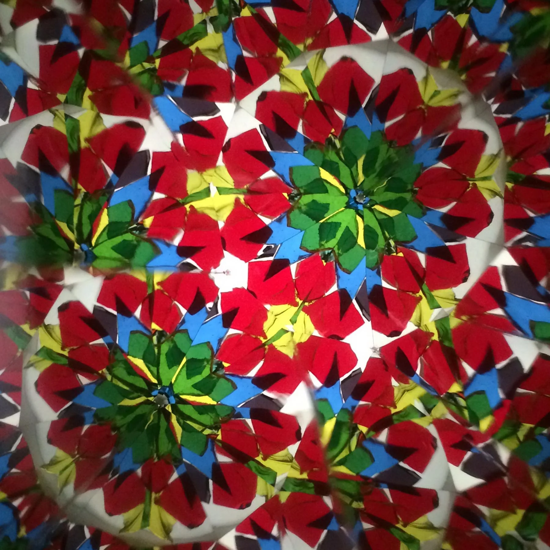  Colorful kaleidoscope pattern showing small blue, red, and green glass flowers clustered together in a complex, blooming geometric arrangement. 