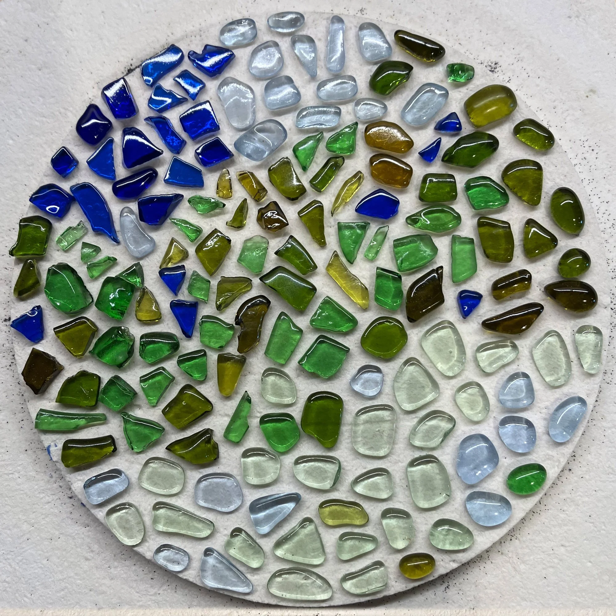  Close-up of arranged recycled glass fragments ready for fusing in a microwave kiln, featuring shades of green and blue. 
