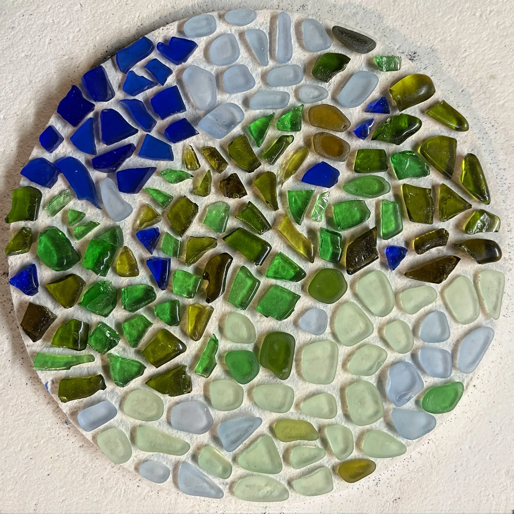  Circular mosaic of colorful recycled glass shards including emerald green, cobalt blue, and amber on a white textured background. 