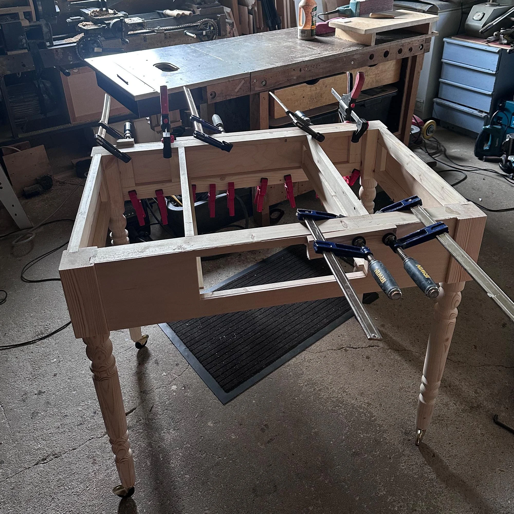  Unfinished wooden desk frame being assembled in a workshop, clamped with blue clamps, visible turned legs. 