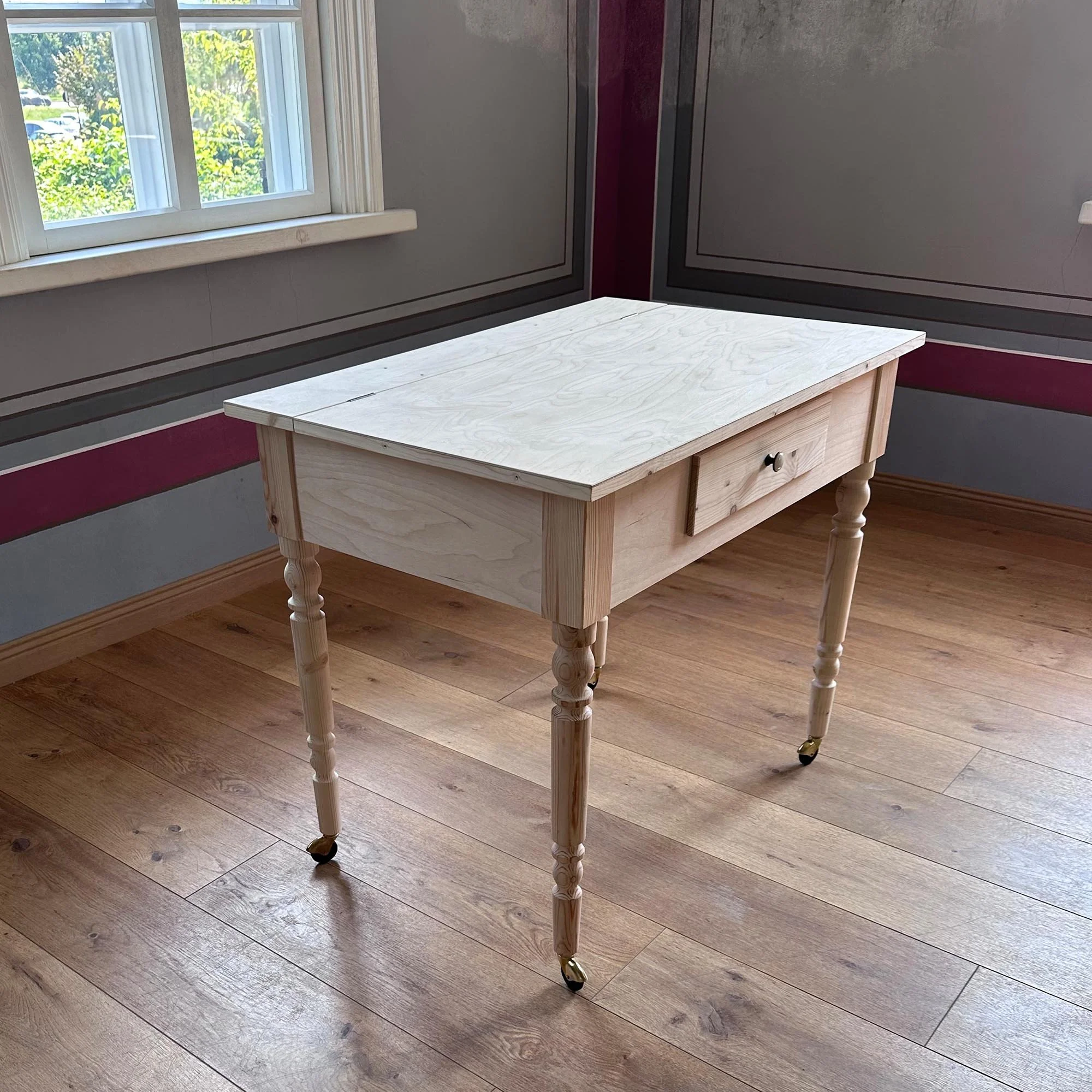  Finished, light-wood antique-style writing desk standing in a sunny room with hardwood floors. 