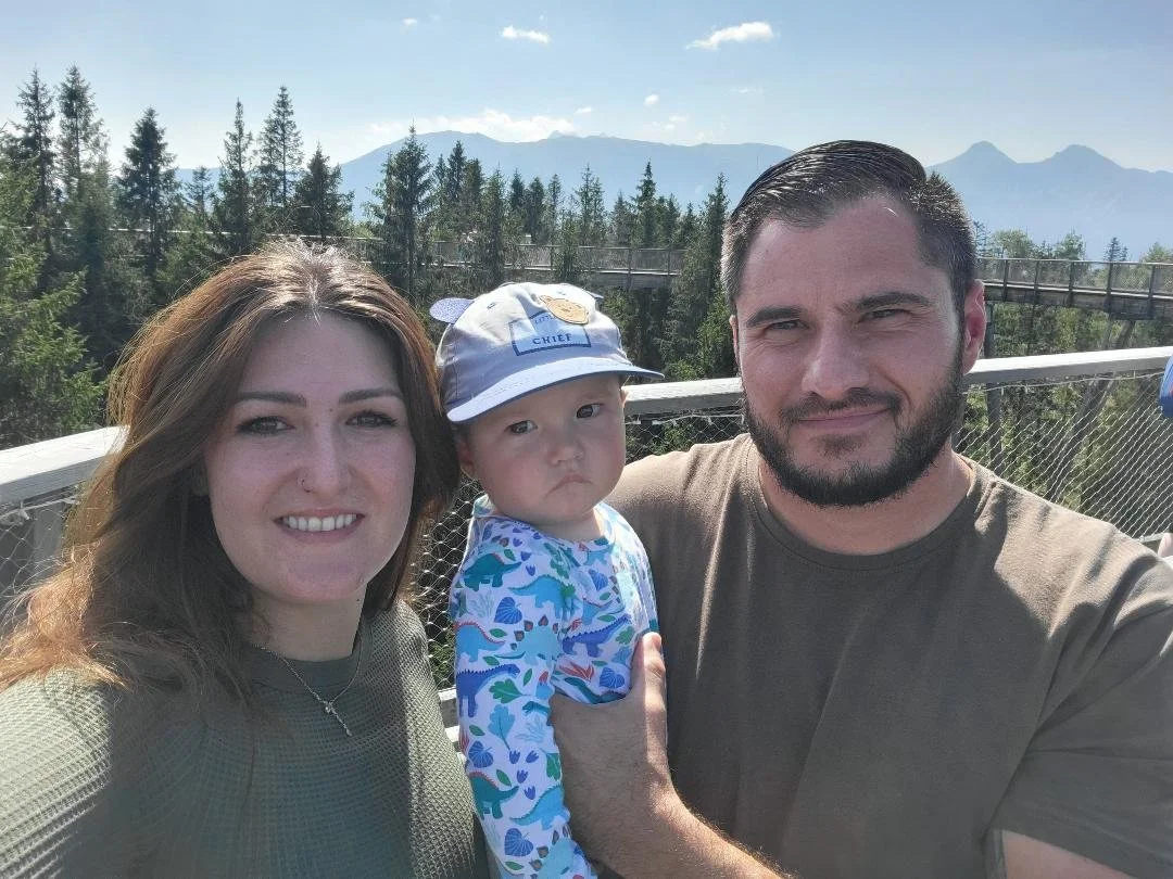 Family of three taking a selfie outdoors with trees and mountains in the background, under a partly cloudy sky.