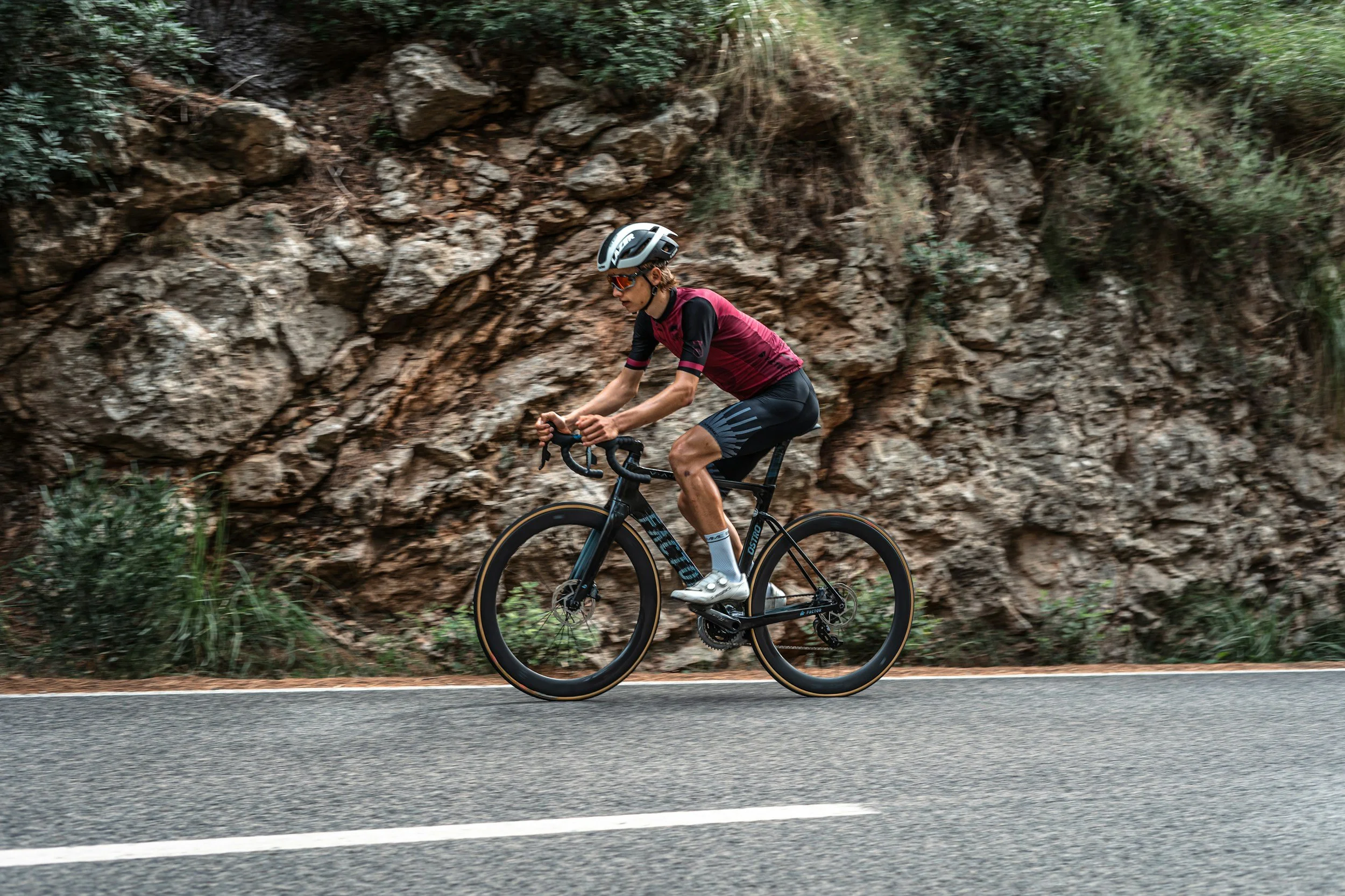 A person riding a black road bike on a paved road next to a rocky hillside with green plants, wearing a helmet and sunglasses.