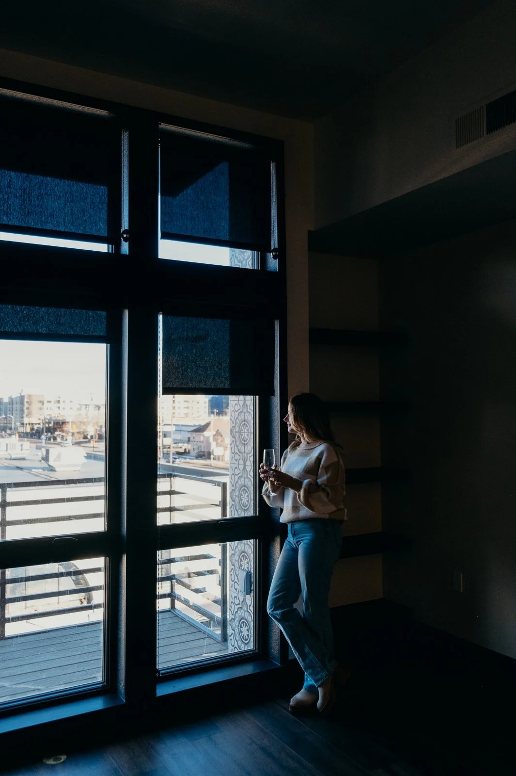 A backlit woman looks out at the city, holding a wine glass