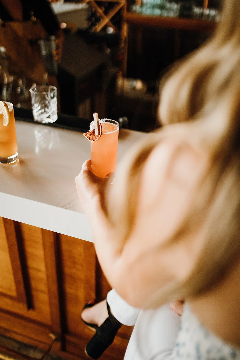 A woman grasps an orange drink on a bartop