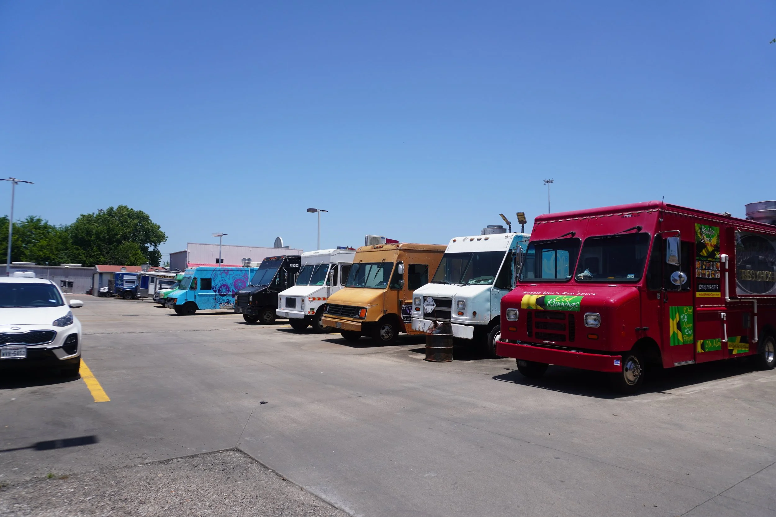 Parking lot with a variety of food trucks, some with colorful designs, and a few parked cars under a clear blue sky.