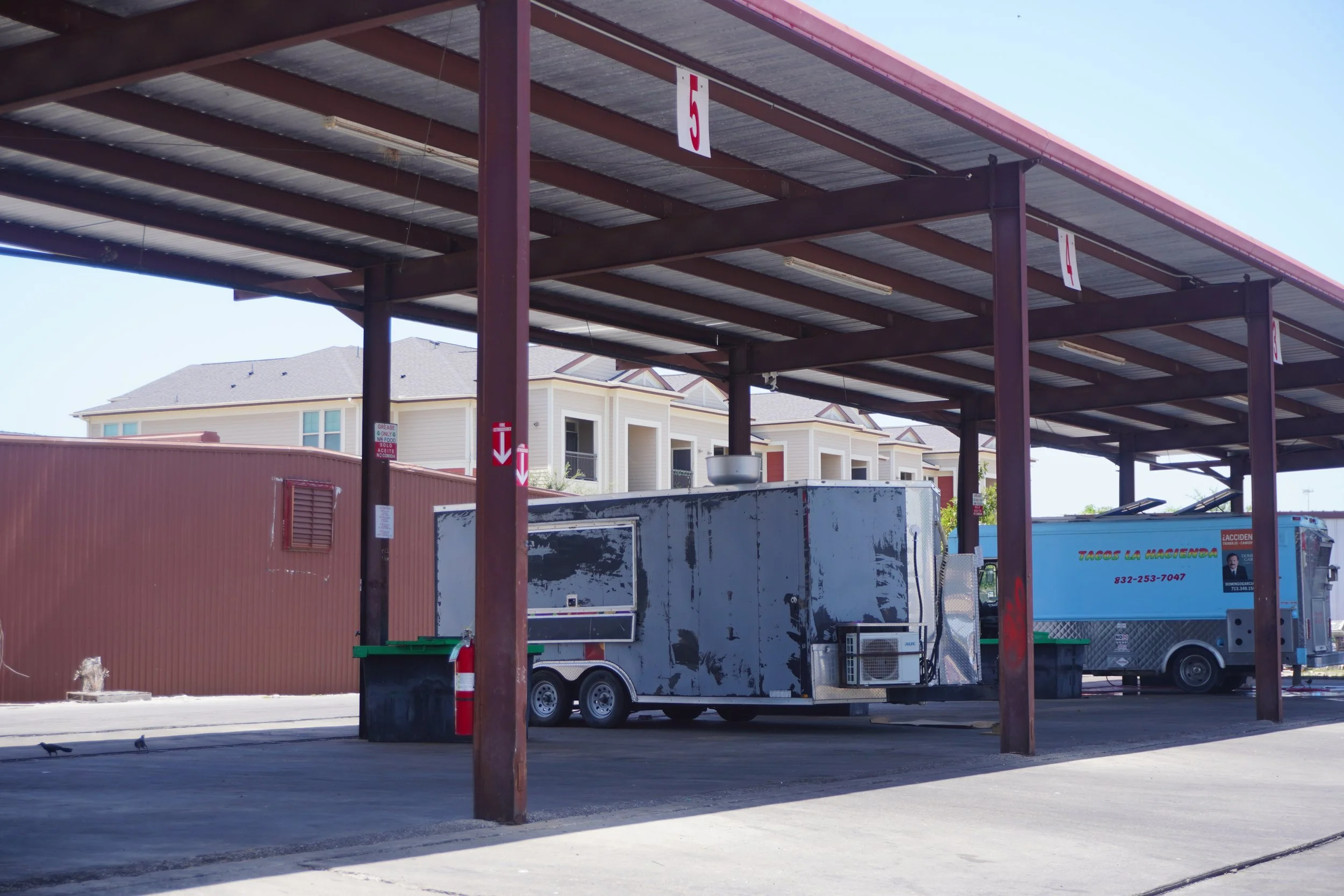 Empty carport with a metal roof, supported by brown metal beams, over a trailer and food truck, with residential buildings in the background.