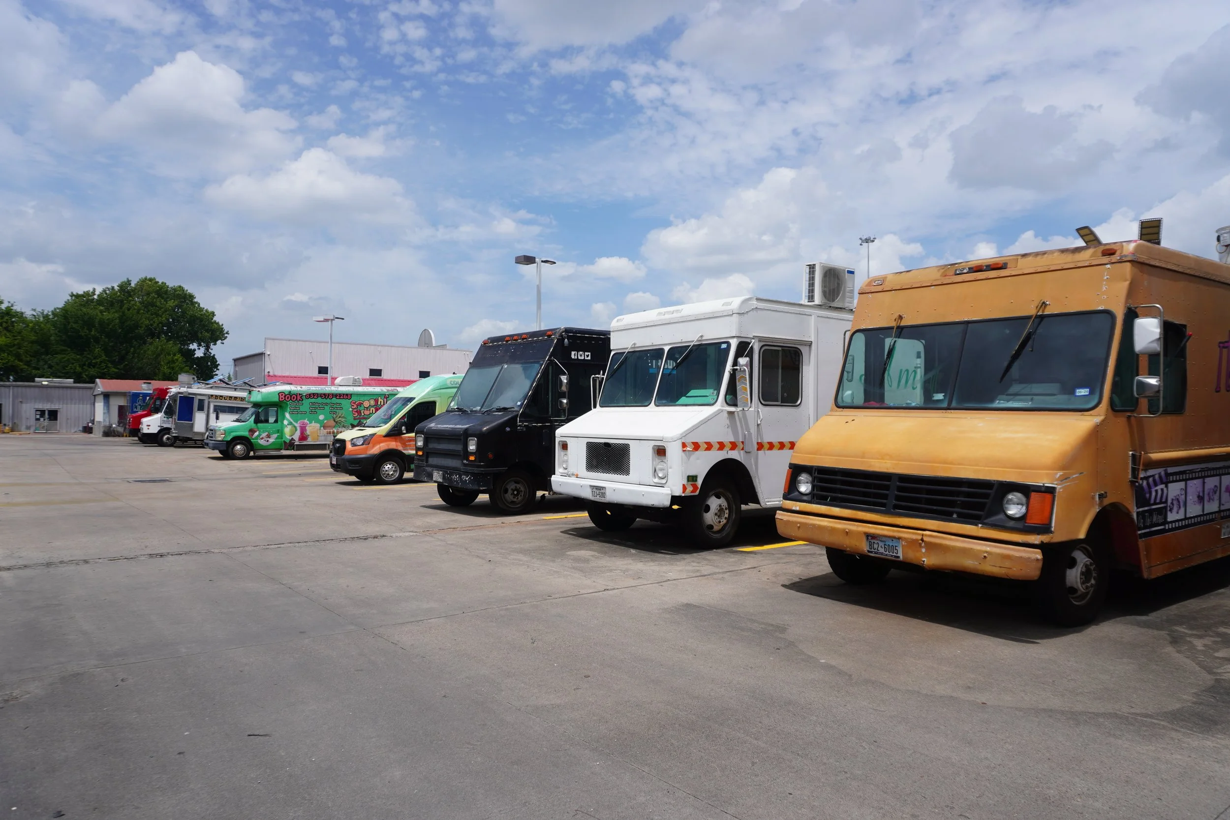 A parking lot with food trucks, including a black truck with Facebook icons, a green smoothie truck, a white truck, a red truck, and a yellow truck, under partly cloudy skies.