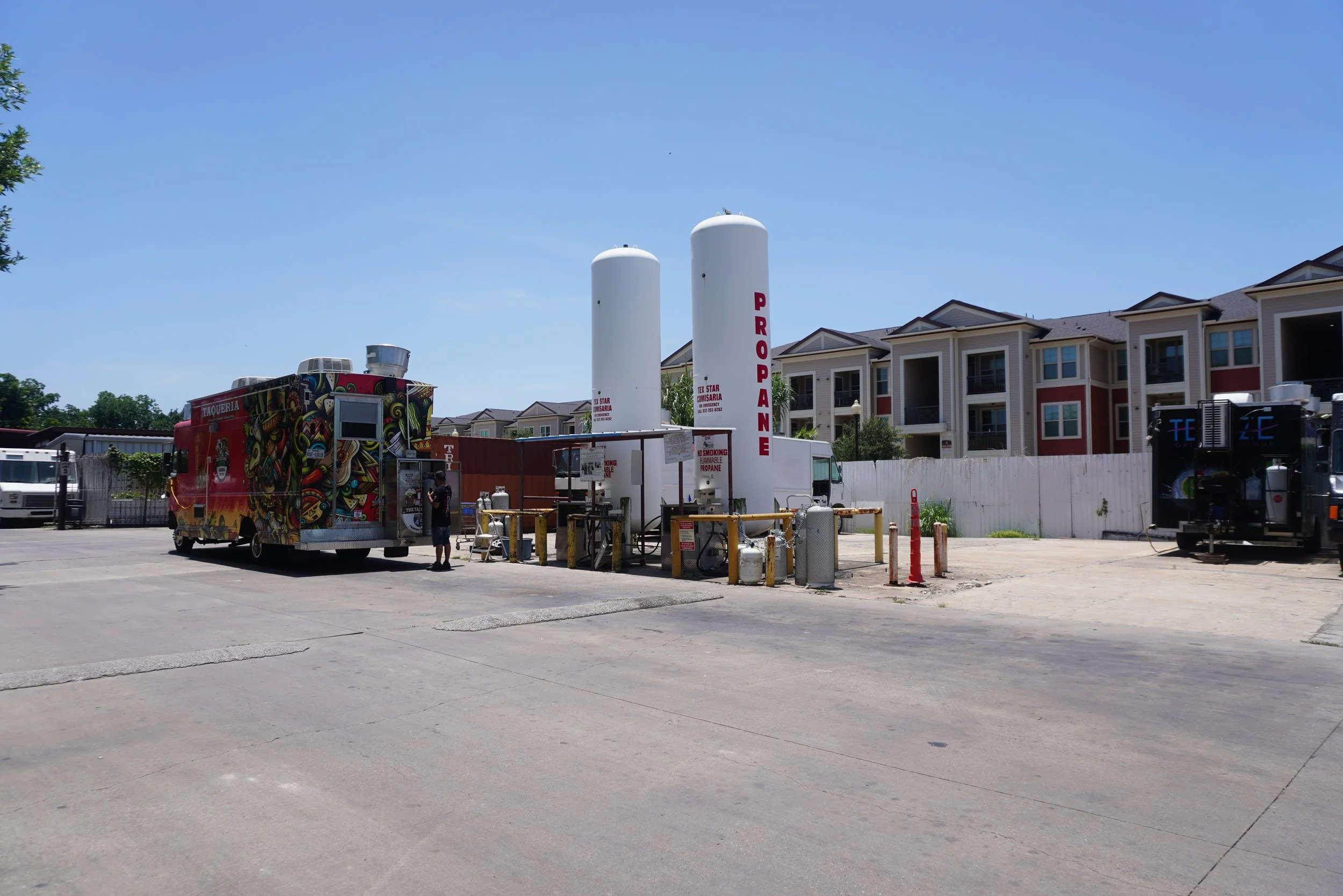A propane filling station with two large vertical propane tanks and a colorful food truck parked nearby. In the background, there are multi-story residential buildings and a clear blue sky.