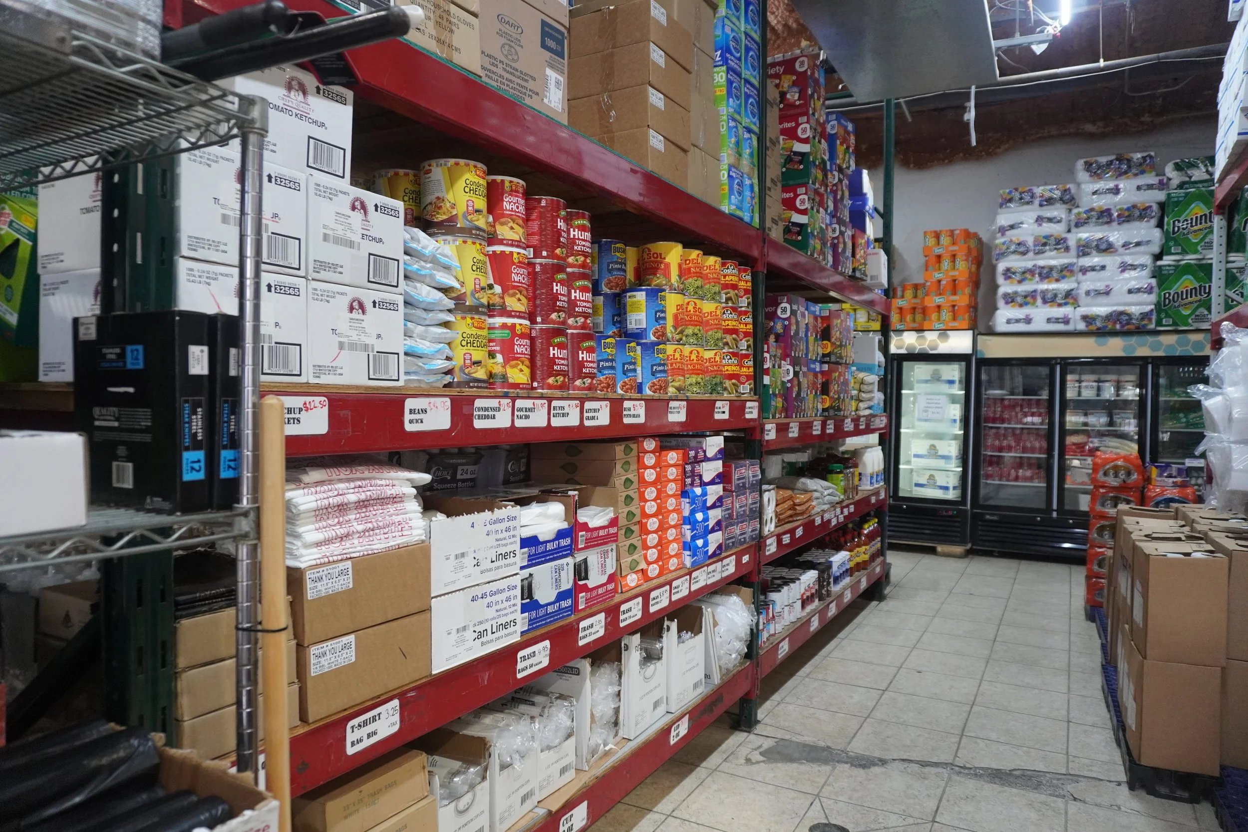 A grocery store aisle with metal shelves stocked with boxes of food products, canned goods, and household supplies, with an aisle extending into the distance and a cooler unit to the right.