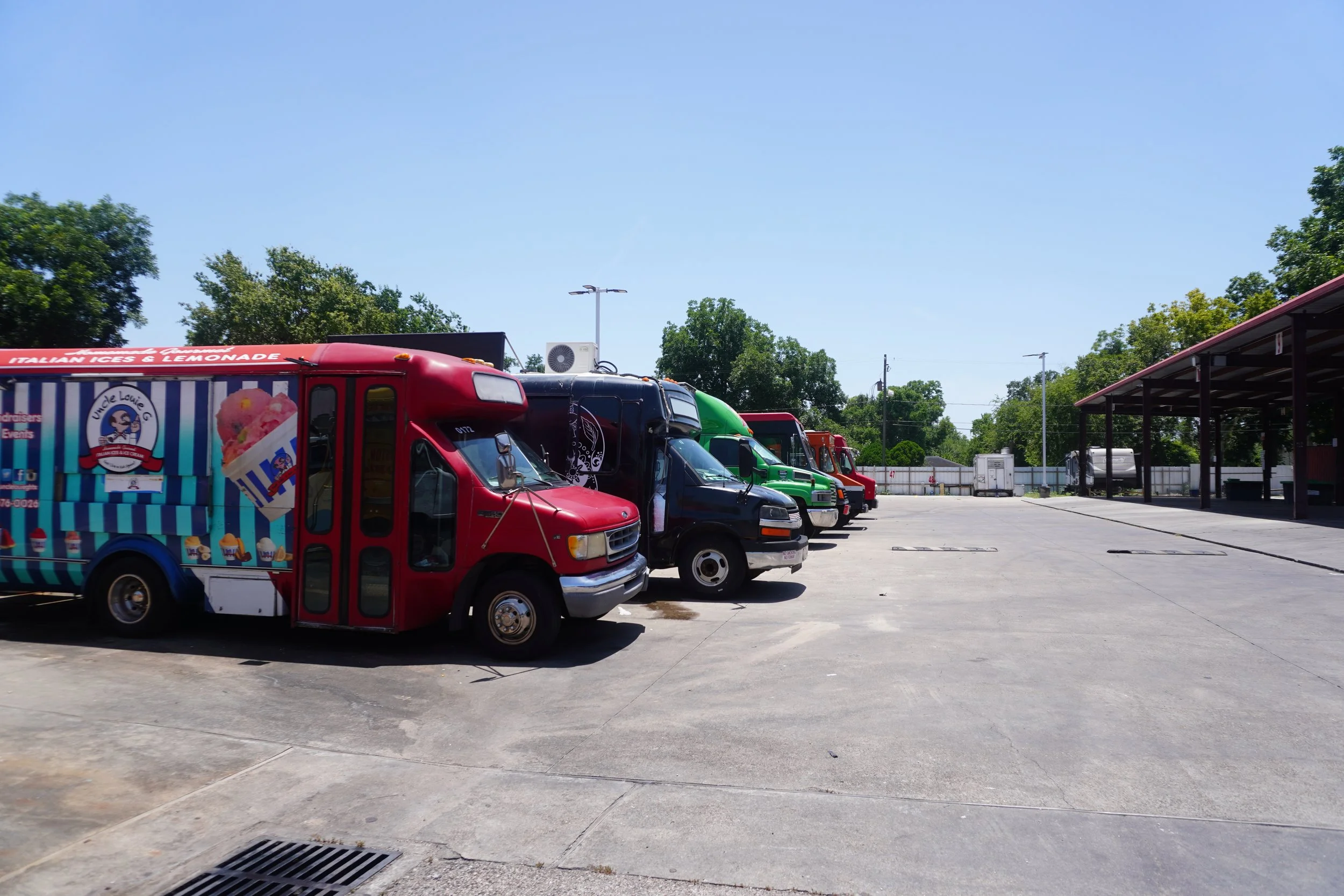Line of food trucks parked in a lot with a covered area and trees in the background.