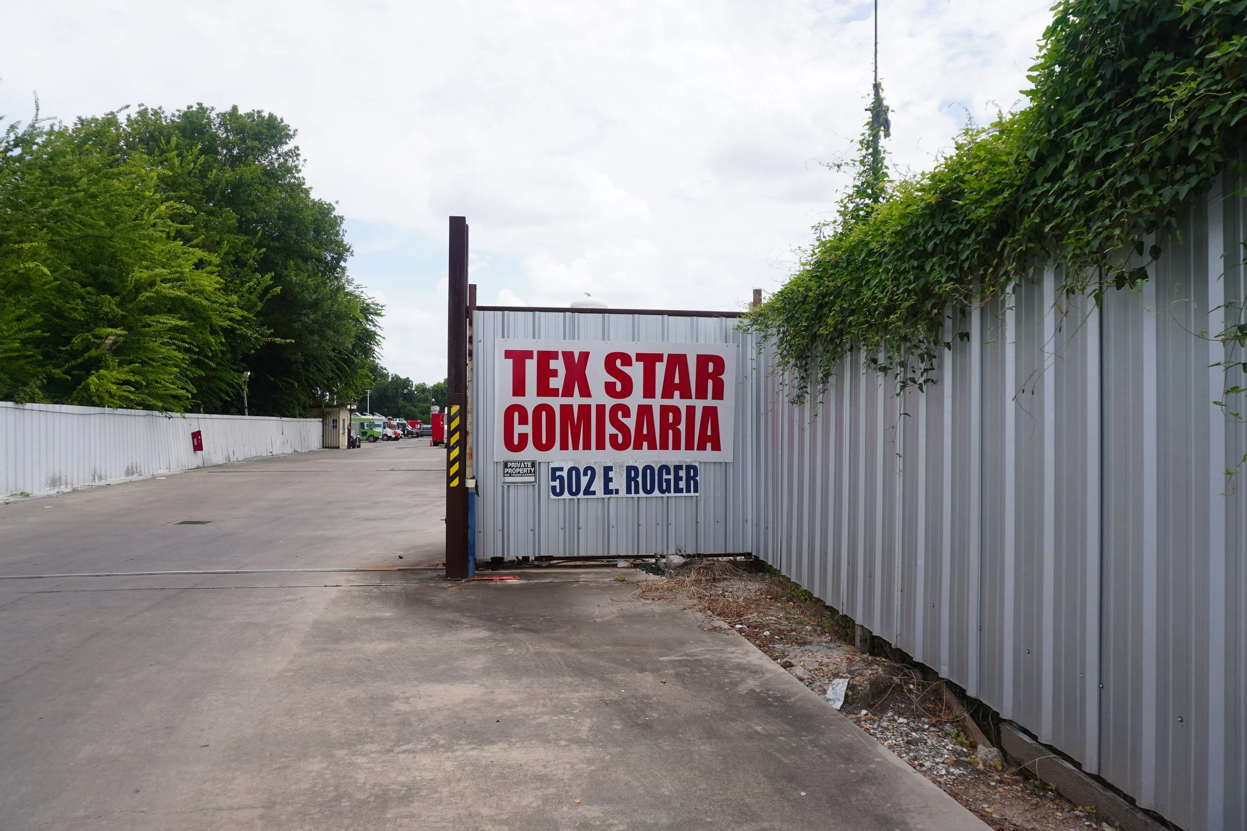 Entrance gate with a sign that reads 'TEX STAR COMISARIA' and an address '502 E. ROGER'. The gate is made of metal panels, and there are cars and a small booth in the background. Green trees are visible on the left side.